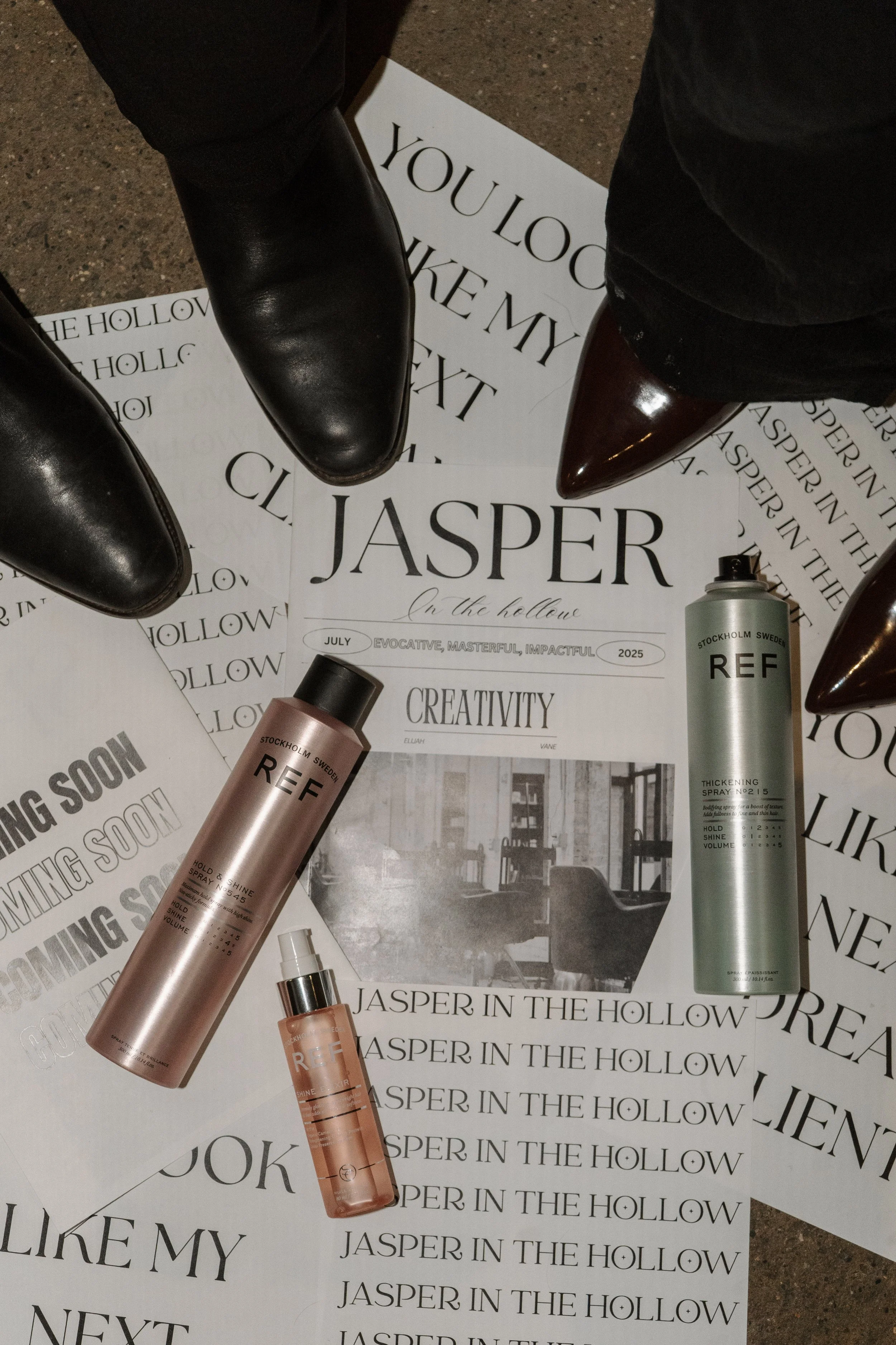 A pair of black leather shoes and three bottles of hair spray on top of printed papers and flyers about Jasper in the Hollow, with a black and white photograph of a living room interior.