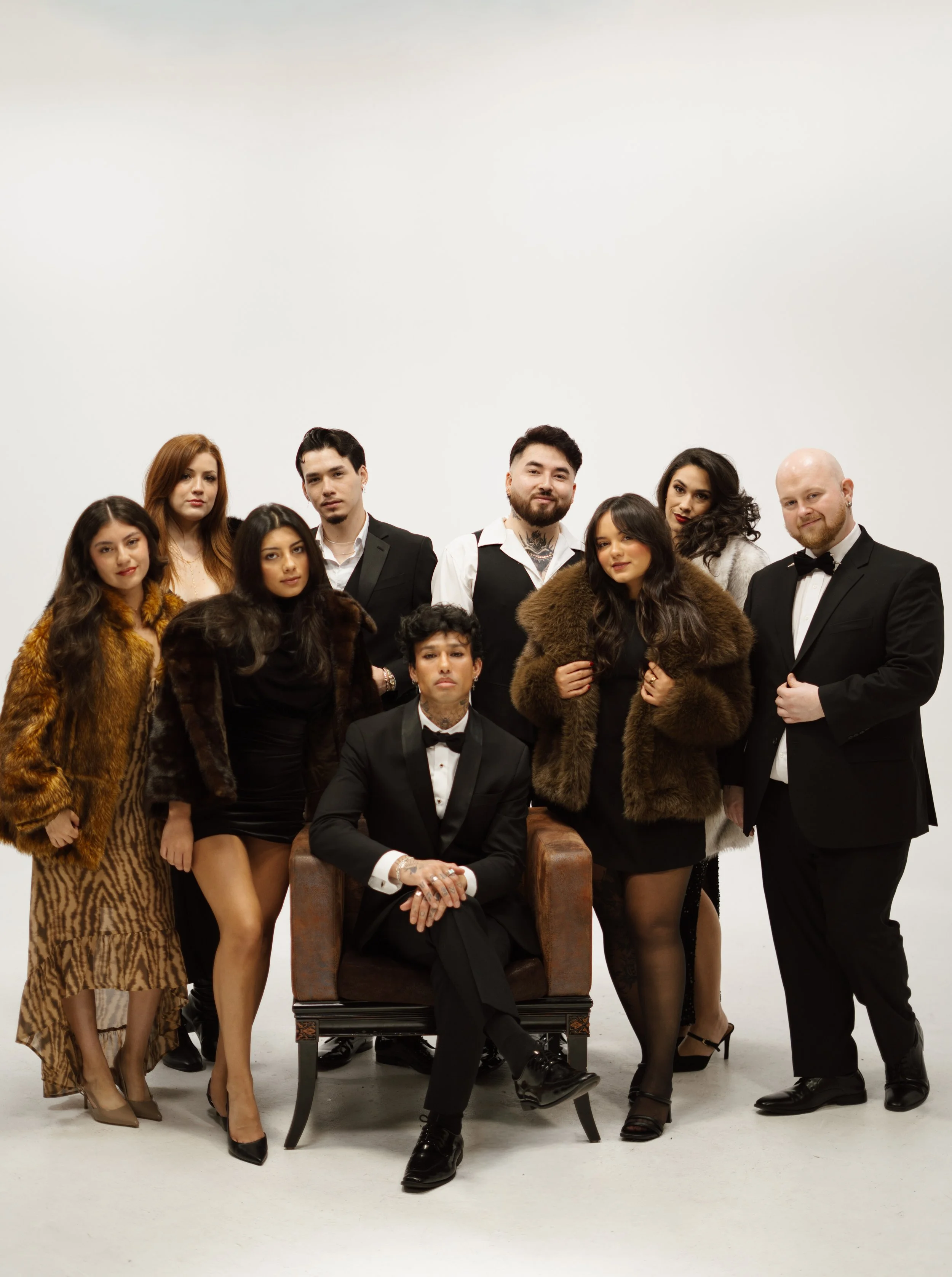 Group of seven diverse people dressed in formal and fashionable outfits posing against a plain white background, with one person seated in a chair.