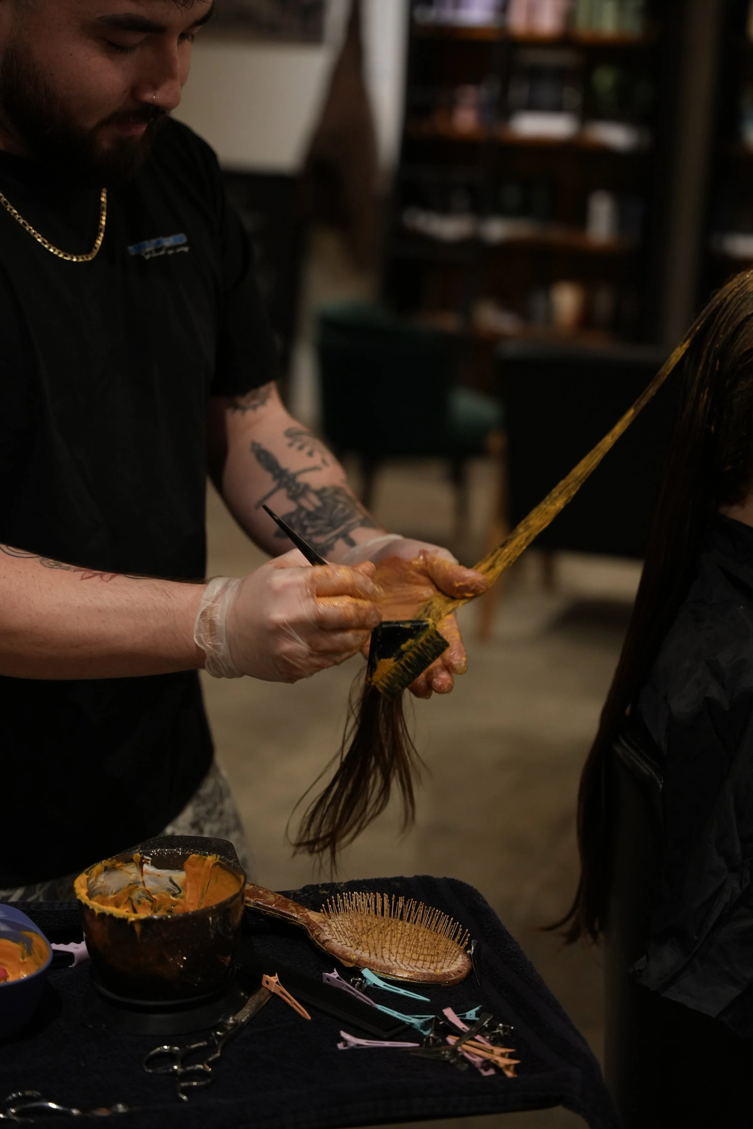 A hairstylist applying hair dye to a woman's long, dark hair in a salon. The stylist is wearing gloves and has tattoos on his arm. There are hairdressing tools and bowls with hair dye on the table.