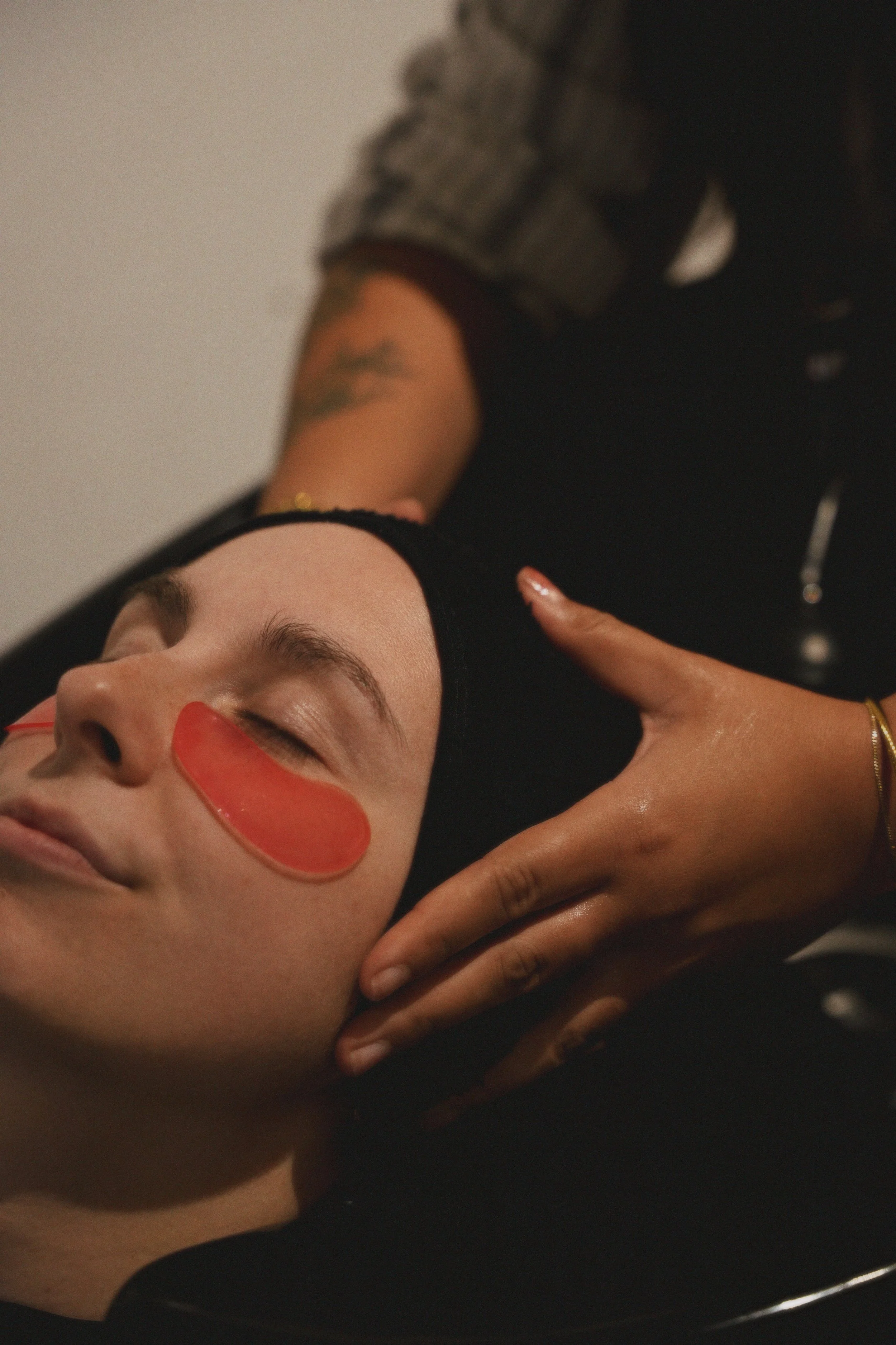 Close-up of a person receiving a facial massage, with a facial mask applied, lying down with eyes closed, and someone gently touching their face.