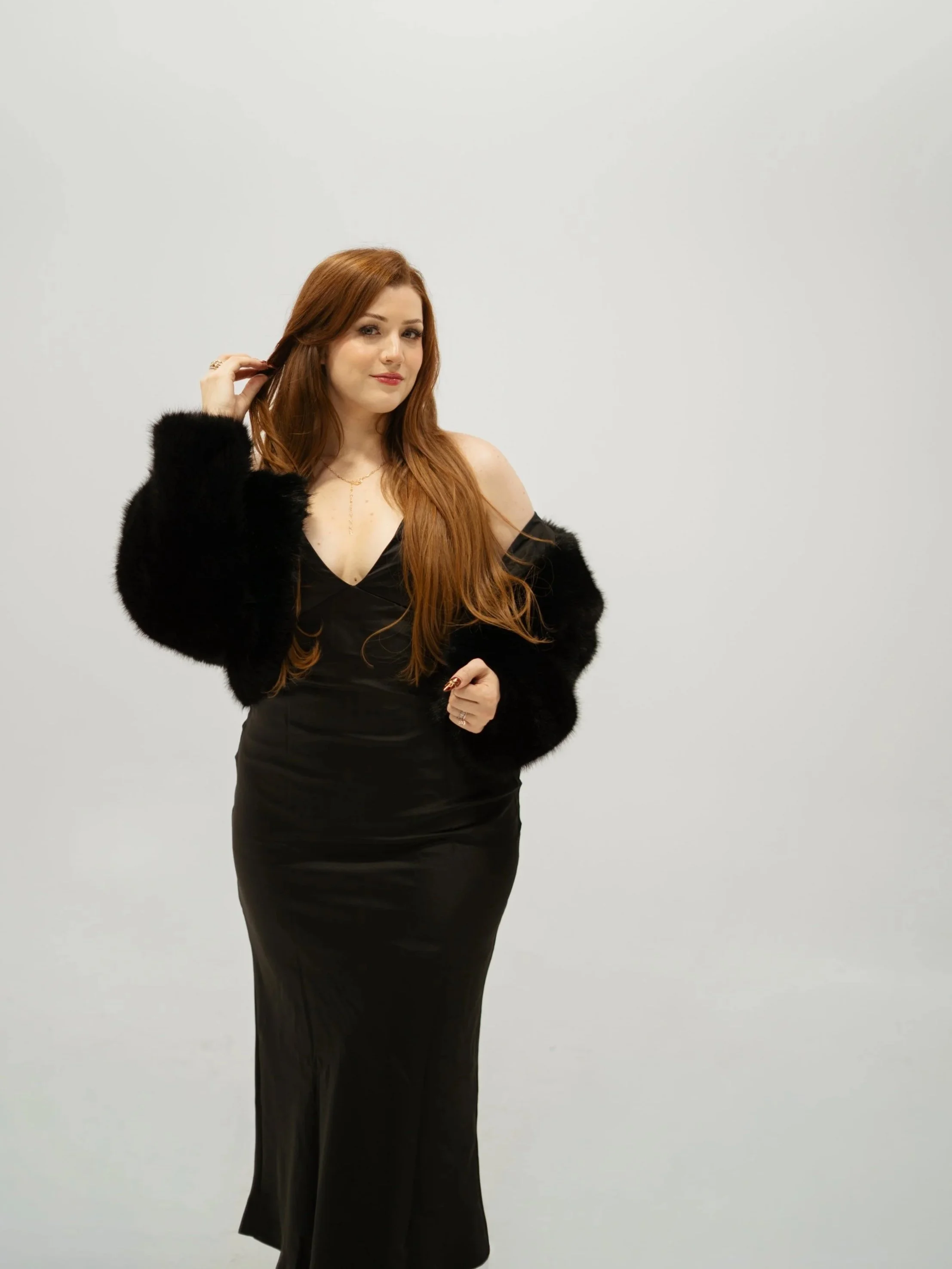 A woman with long red hair wearing a black dress and a black fur coat, standing against a plain white background, looking at the camera with a slight smile.