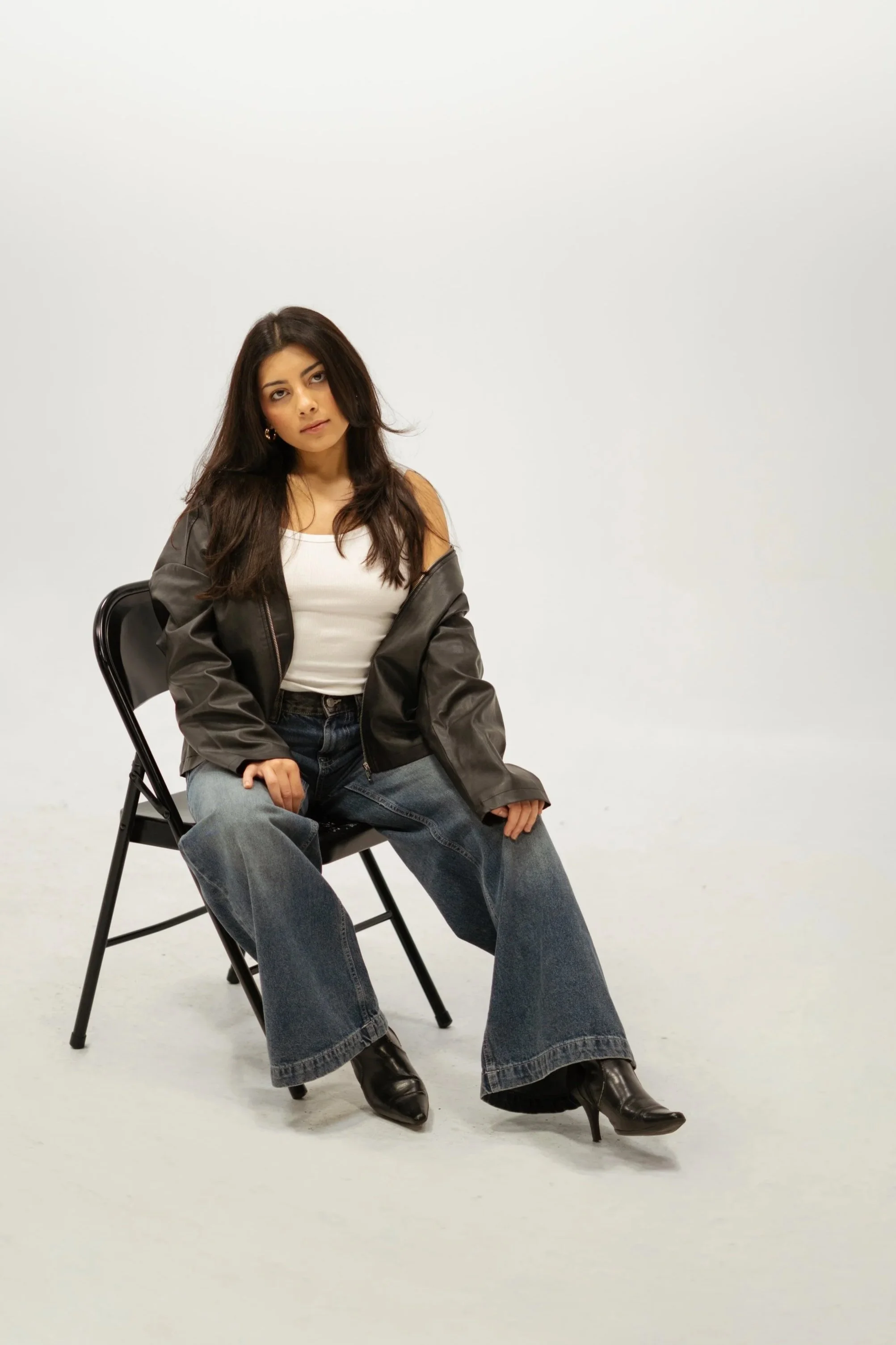 A woman sitting on a black chair against a plain white background. She has long dark hair, wears a white tank top, a black leather jacket, large jeans, and black pointed leather boots with heels. She appears to be posing with her head tilted slightly and a relaxed expression.
