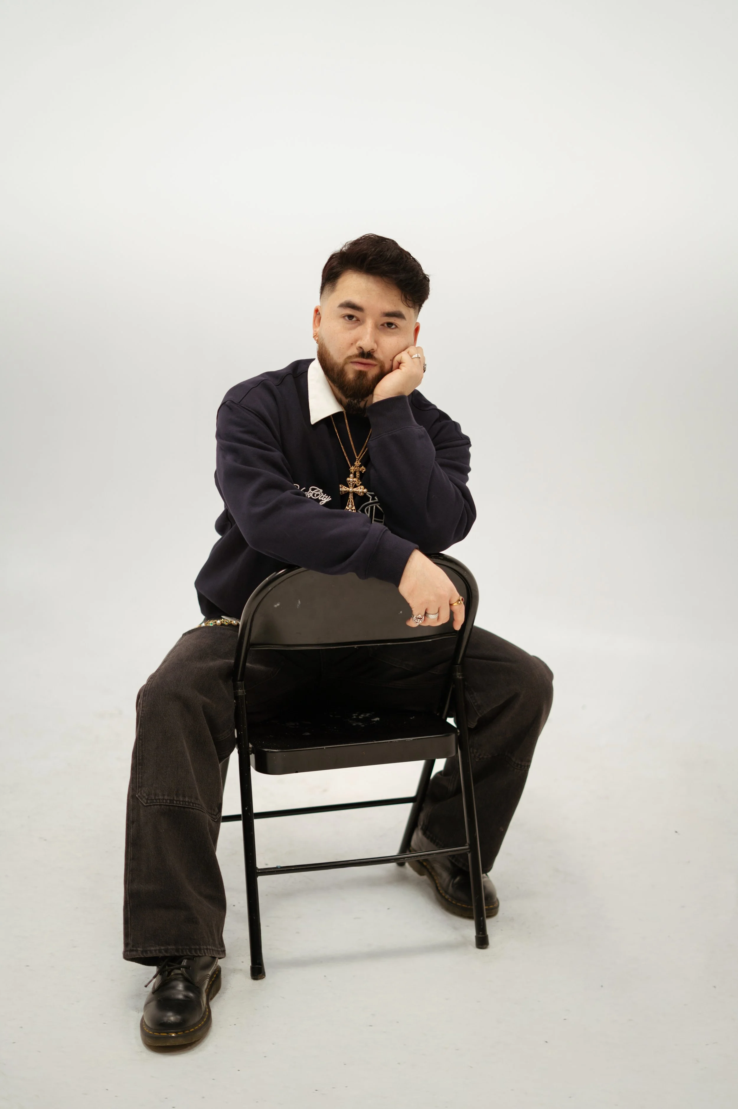 A young man with a beard and short dark hair sitting on a black chair against a plain white background. He is resting his chin on his hand, looking at the camera, wearing a black sweatshirt, dark pants, and black boots. He has jewelry including rings, necklaces, and earrings.