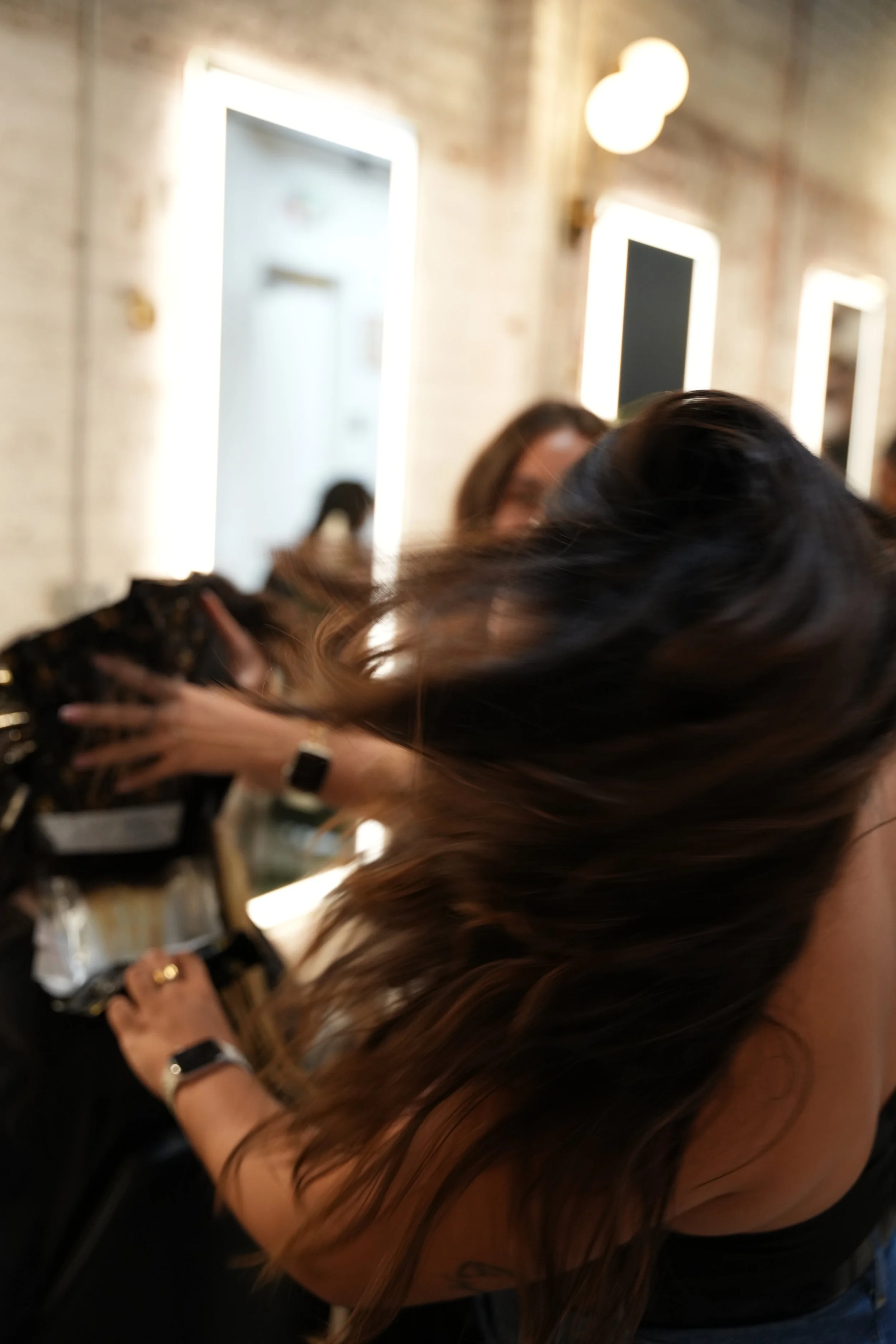 A woman with long, dark hair is sitting indoors, looking at her phone. The background shows a brick wall with illuminated frames and round ceiling lights.