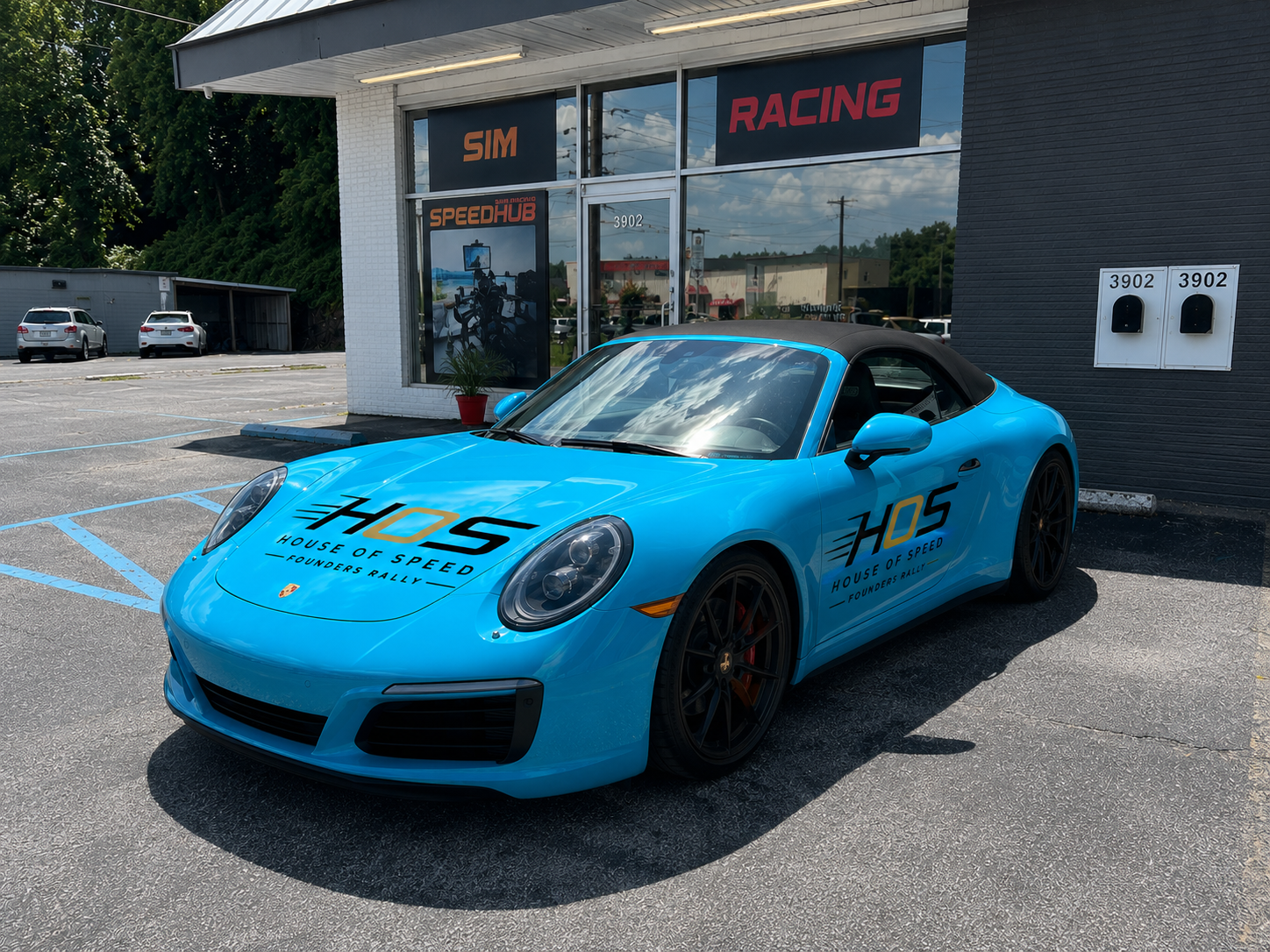 Blue Porsche sports car with 'HOS House of Speed' logo parked outside a racing shop with large windows and signage, including 'SIMA' and 'RACING.'