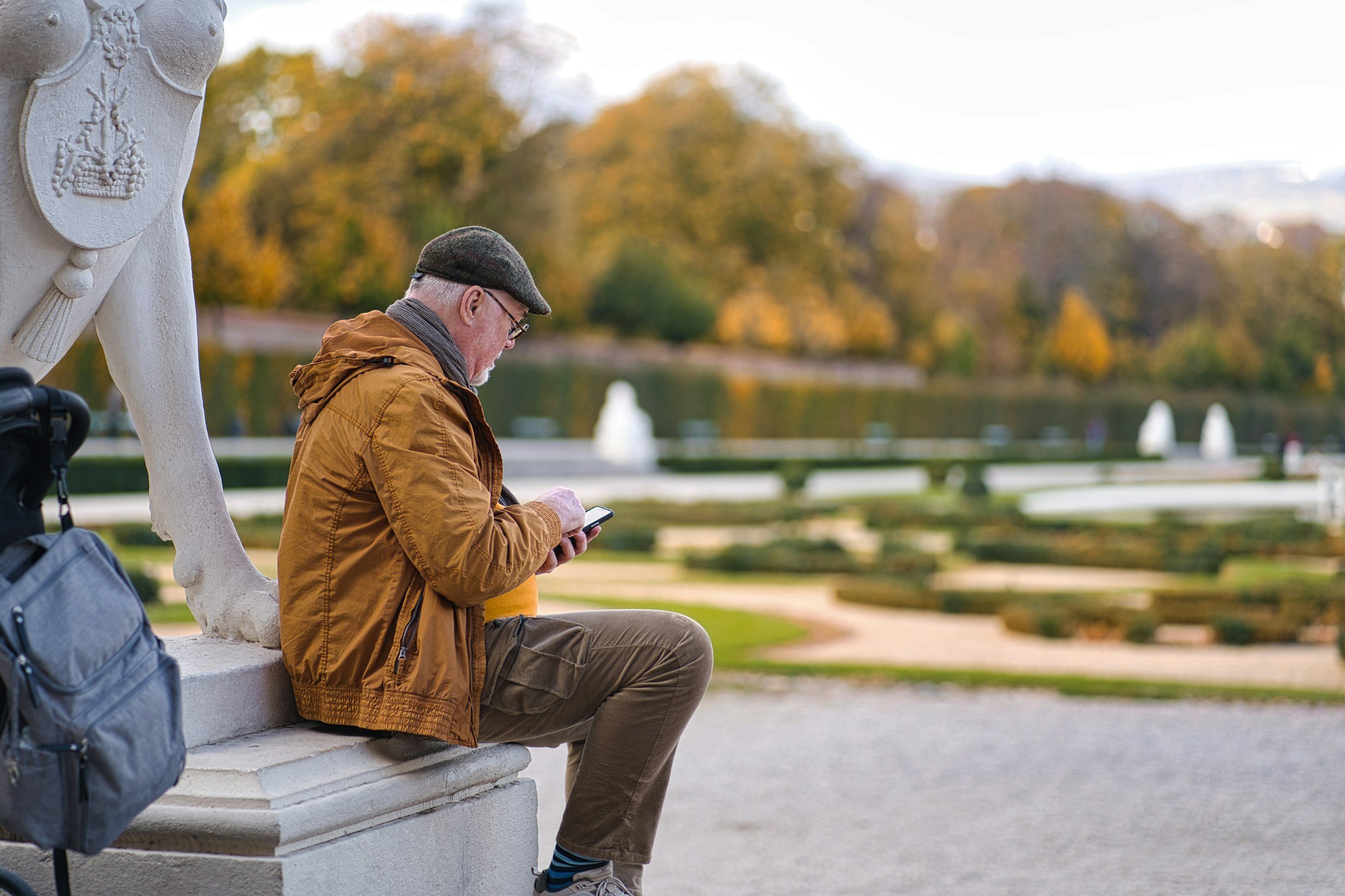 An older man wearing a green beret and sunglasses sits on a park bench, looking at his phone, with a large white sculpture of an animal's leg and a backpack beside him. The background features trees with autumn foliage and a pond with fountains.