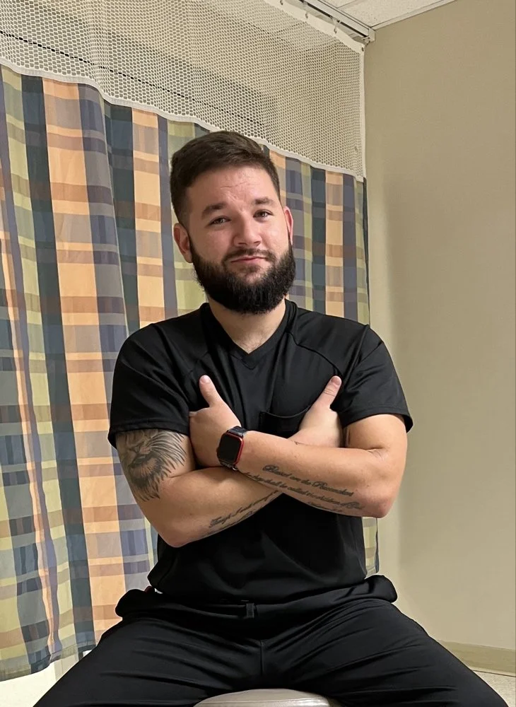 A man with a beard and tattoos on his arms sitting on a stool, crossing his arms and making a playful face, in a room with patterned curtains and a beige wall.