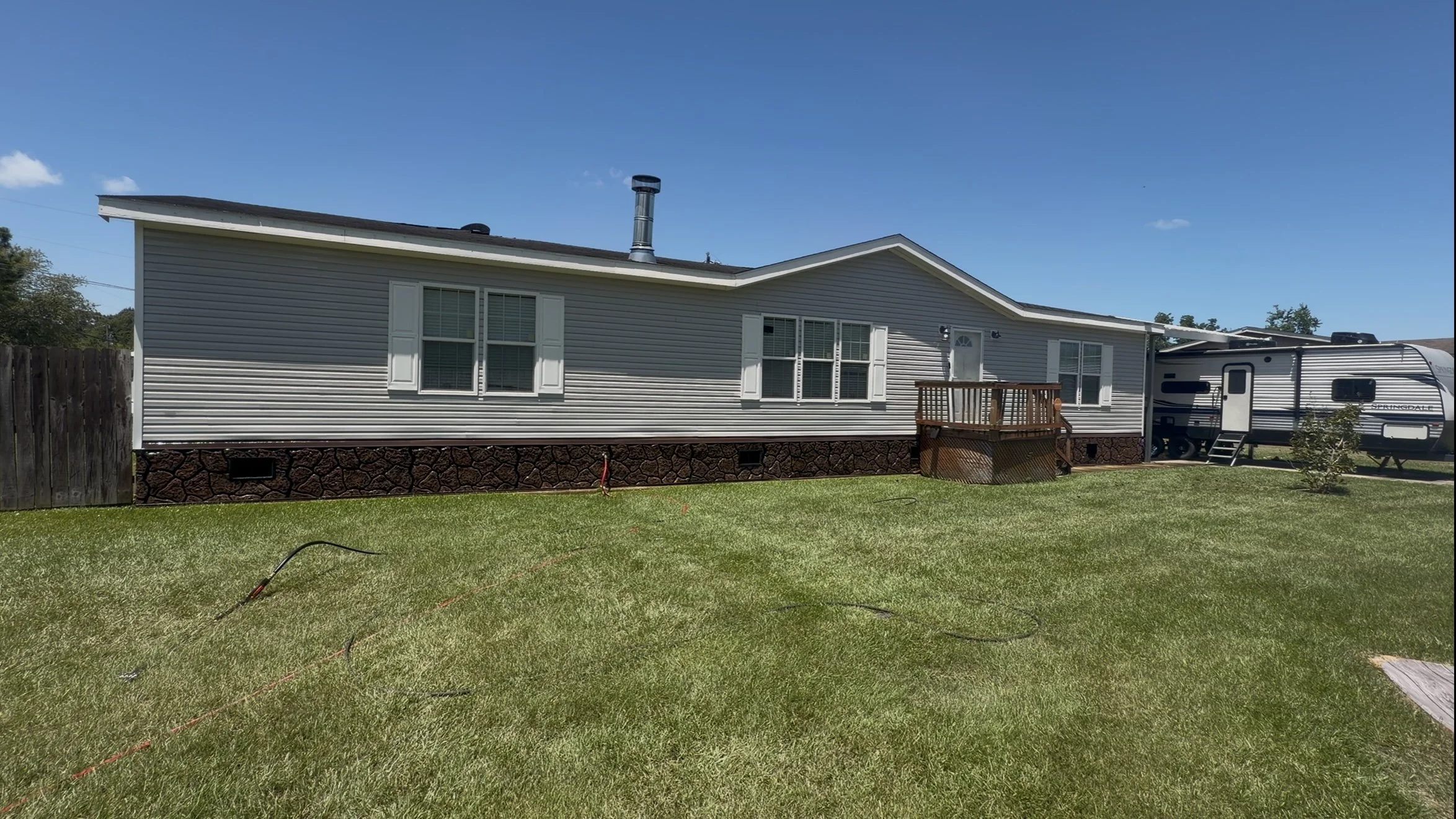 Backyard with a manufactured home, small wooden deck, RV parked on the grass, and a cloudy blue sky.