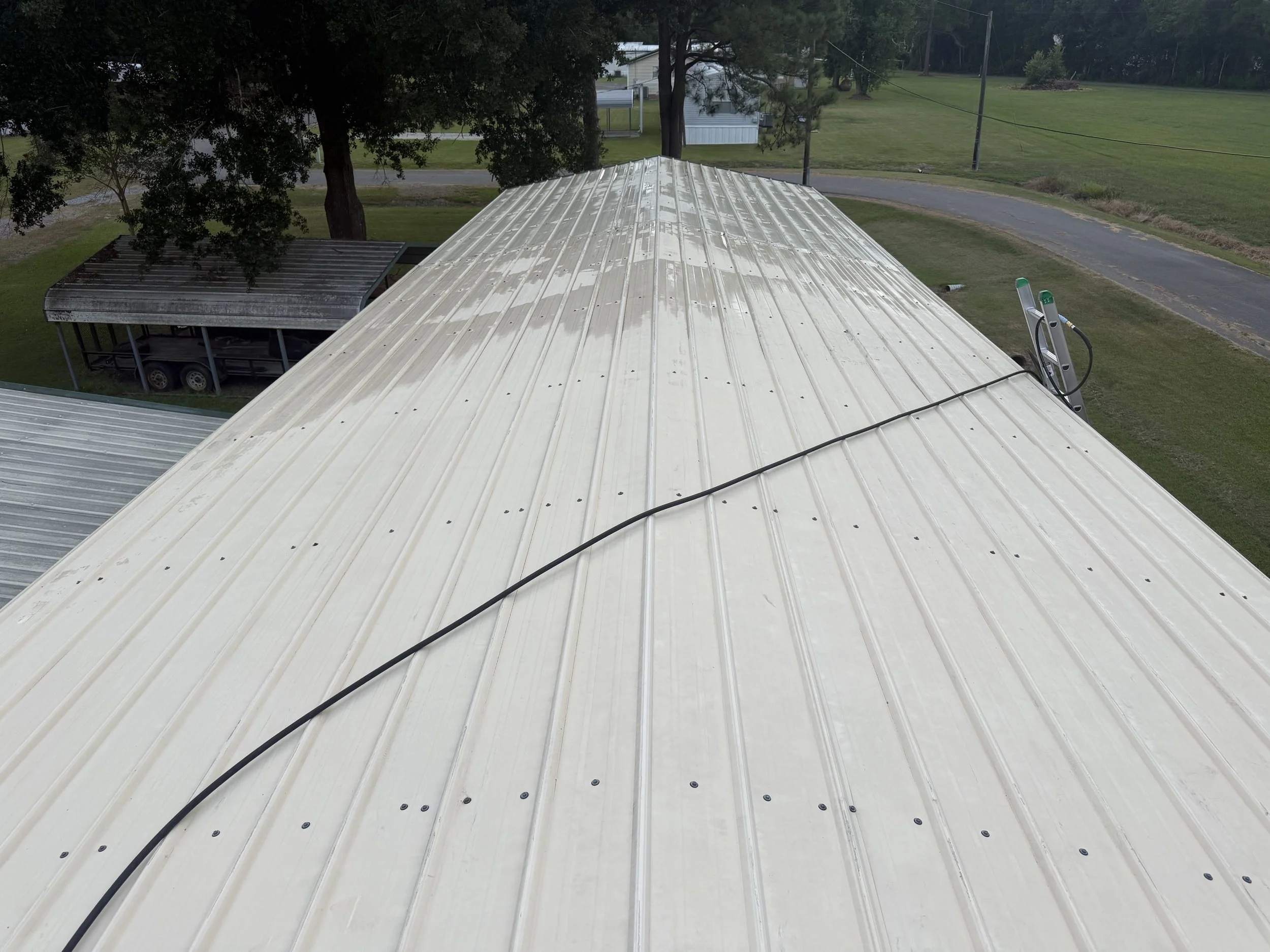 Aerial view of a metal roof with a black power cord or cable running across it, surrounded by green trees, grass, a small road, and utility poles in the background.