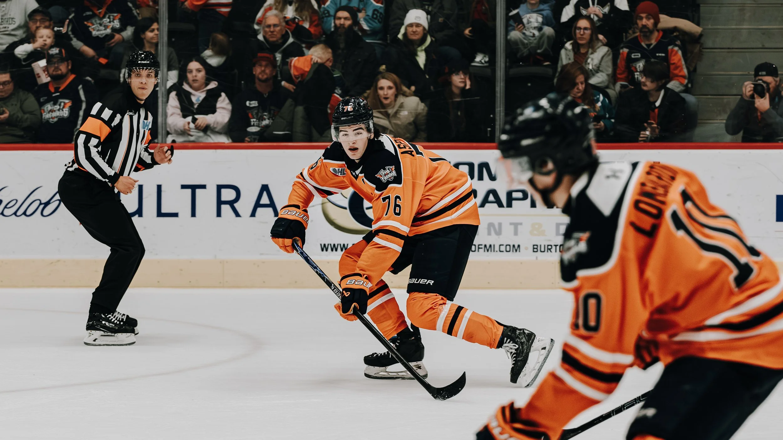 A hockey game with players in orange jerseys and a referee on the ice, with spectators watching from the stands.