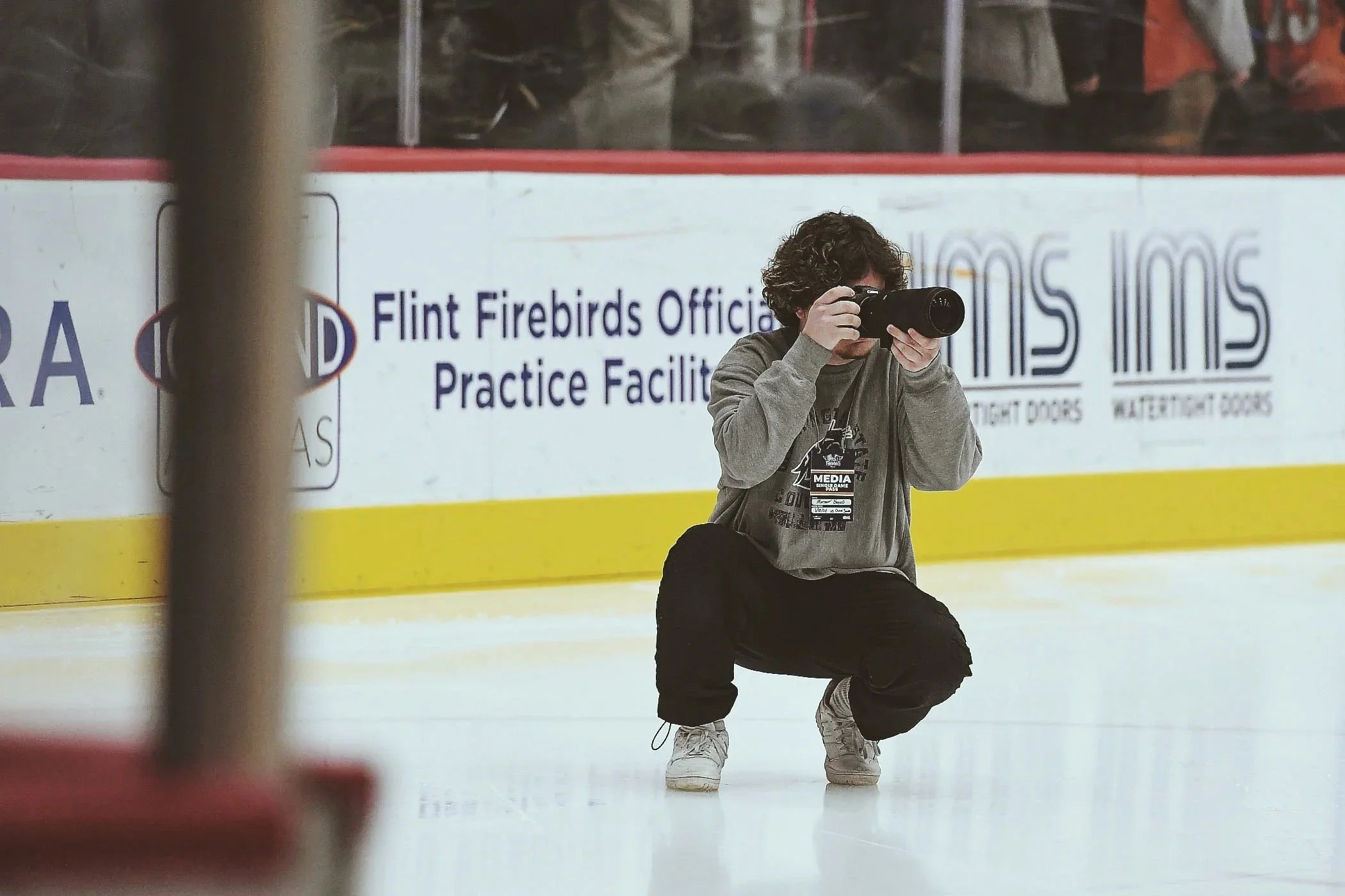 A person kneeling on ice wearing a gray hoodie, black pants, gray and white sneakers, and a media credential around their neck, taking a photograph during a hockey game at an ice rink with a sign for the Flint Firebirds practice facility in the background.