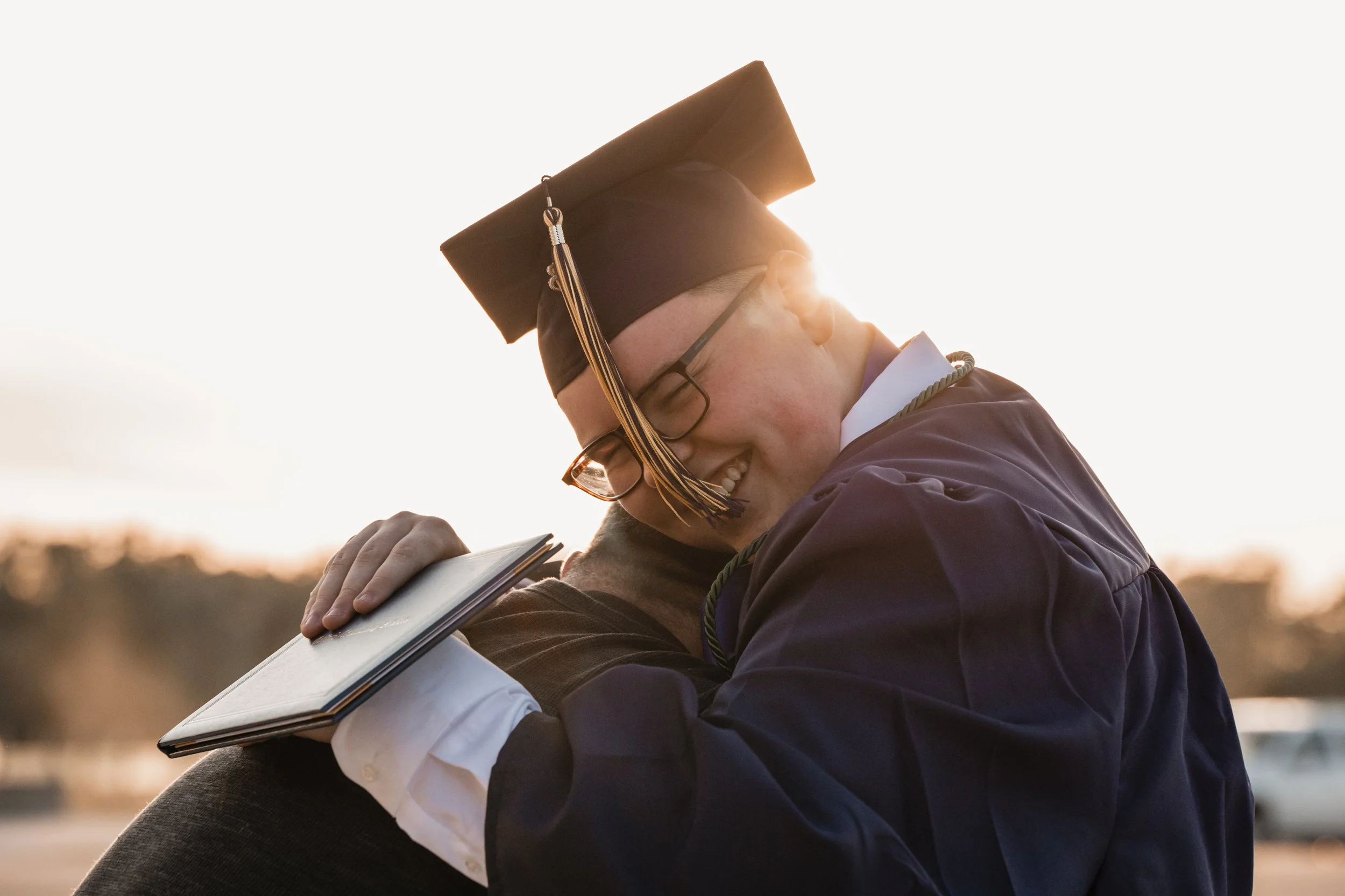 A young man in a graduation cap and gown embraces another person while holding a certificate, with the sun setting in the background.