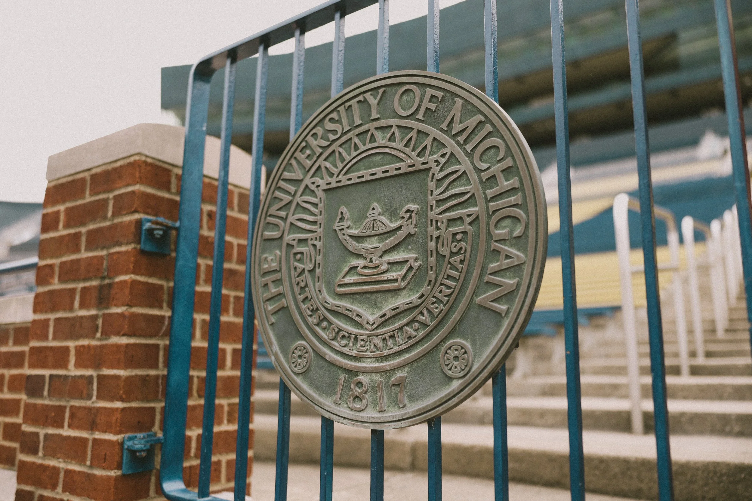 The emblem of the University of Michigan on a metal gate, with a brick pillar on the left and part of a building in the background.