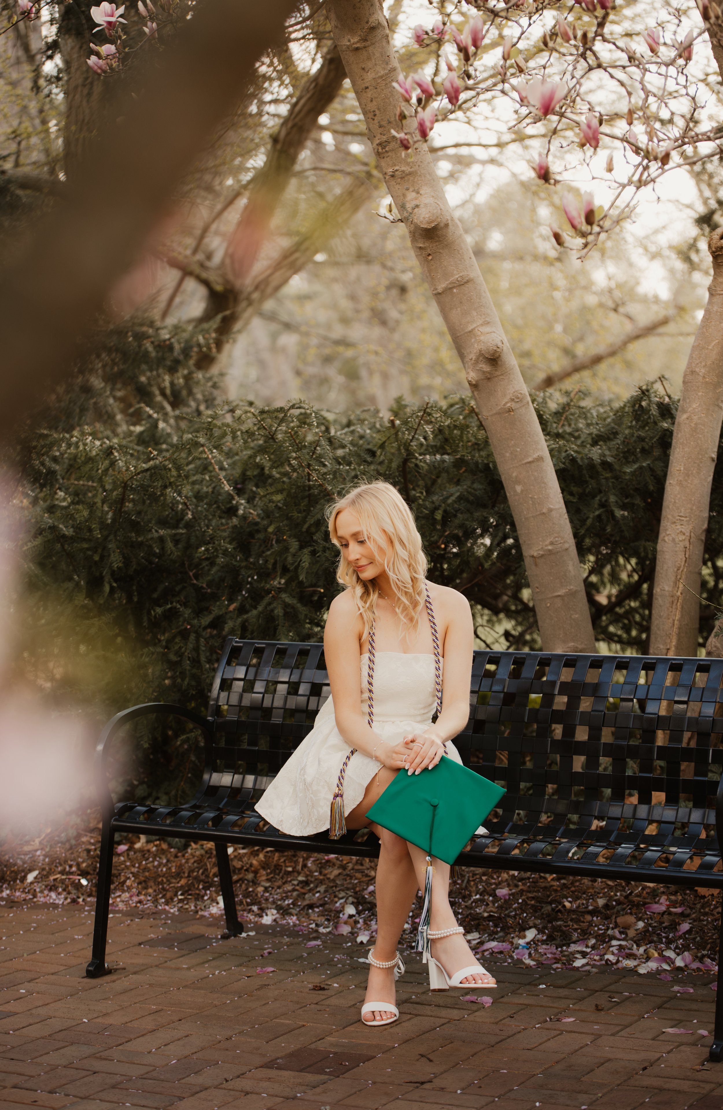 A woman in a white dress sitting on a black metal bench in a park or garden with pink blossoms on trees and a ground covered with pink petals, holding a green handbag and wearing white high-heeled shoes with pearl details.