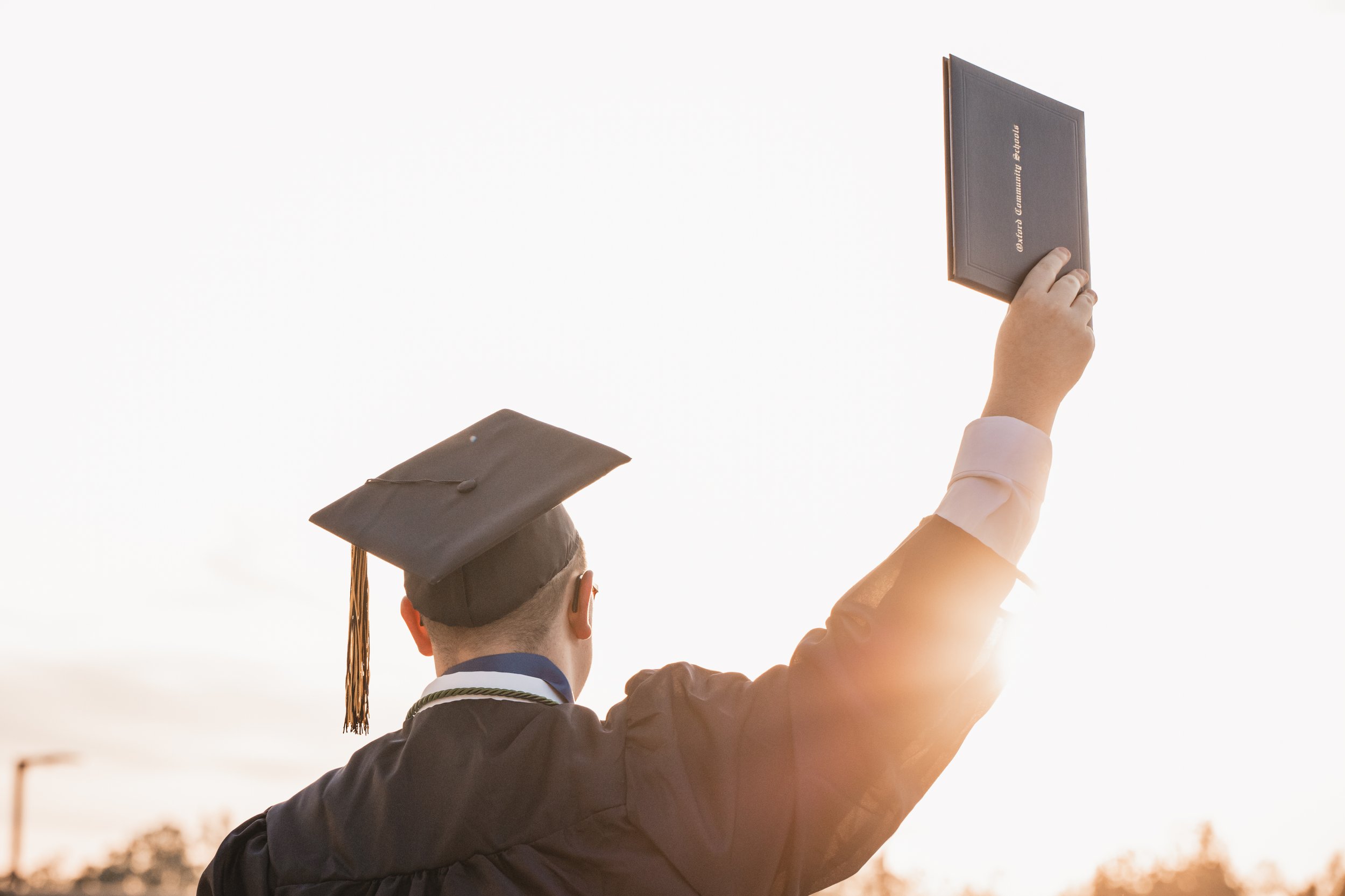 A person in graduation attire holding a diploma in the air during a graduation ceremony outdoors.