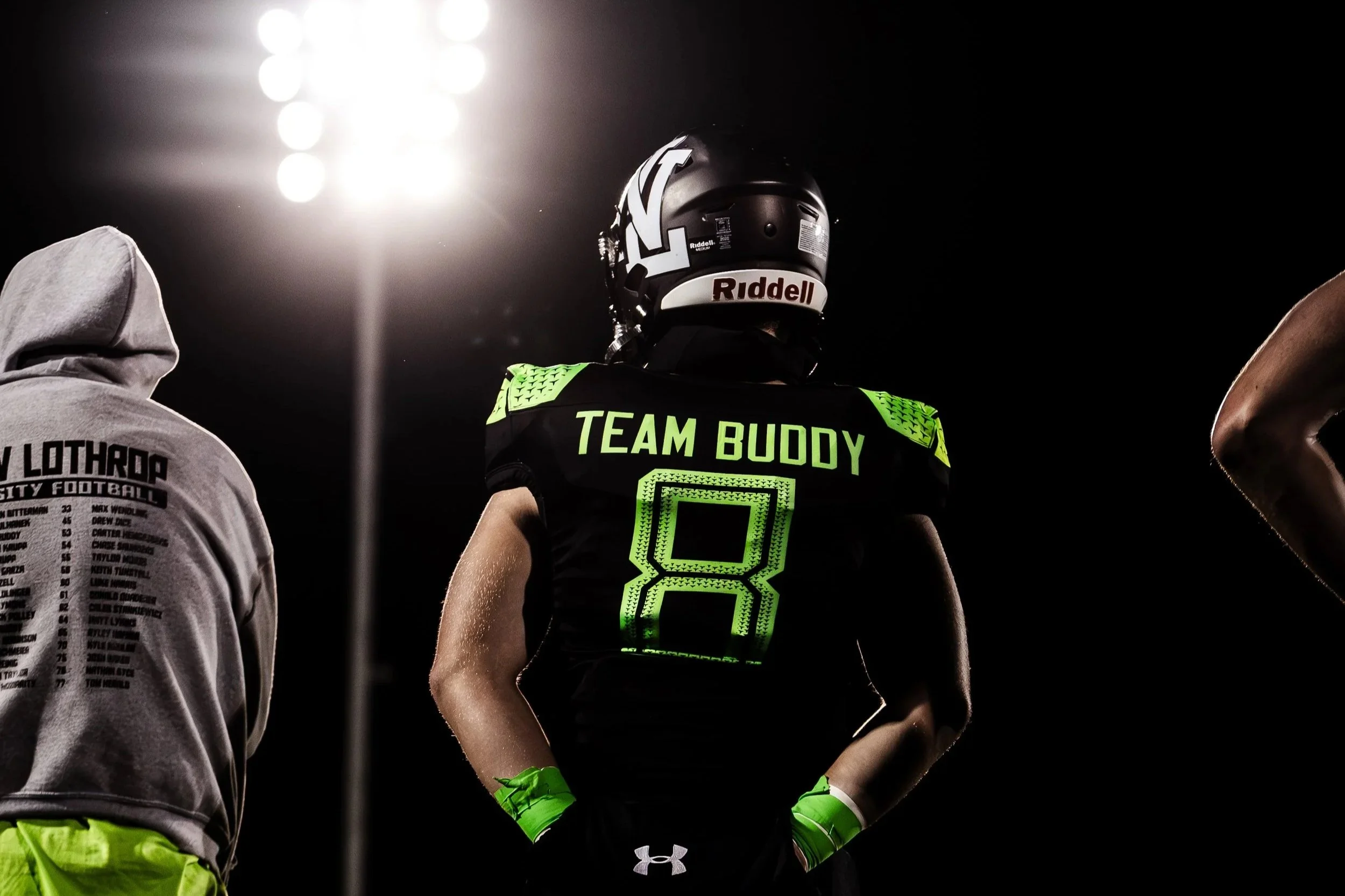 A football player wearing a black helmet and uniform with neon green accents and the words "TEAM BUDDY" on the back, standing on a football field under stadium lights.