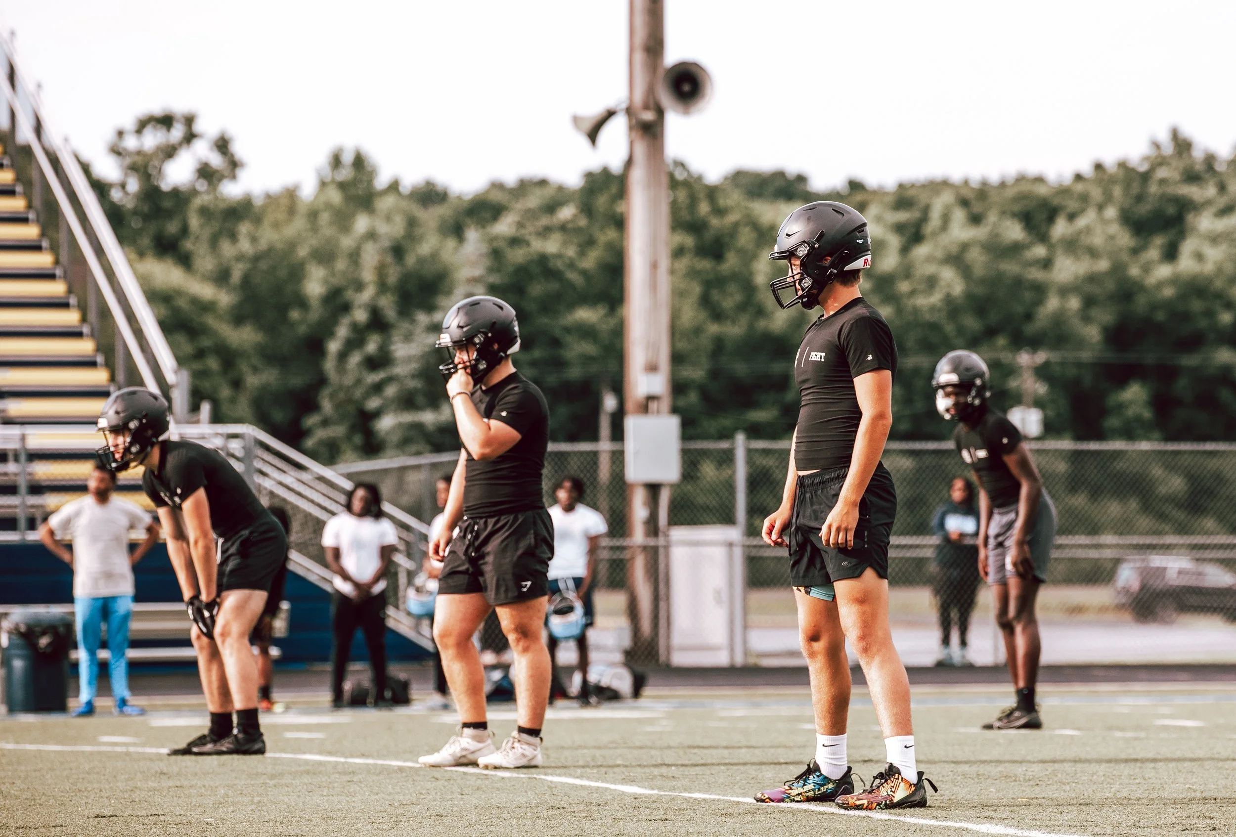 Football players in black helmets and uniforms practicing on a field with people observing in the background.