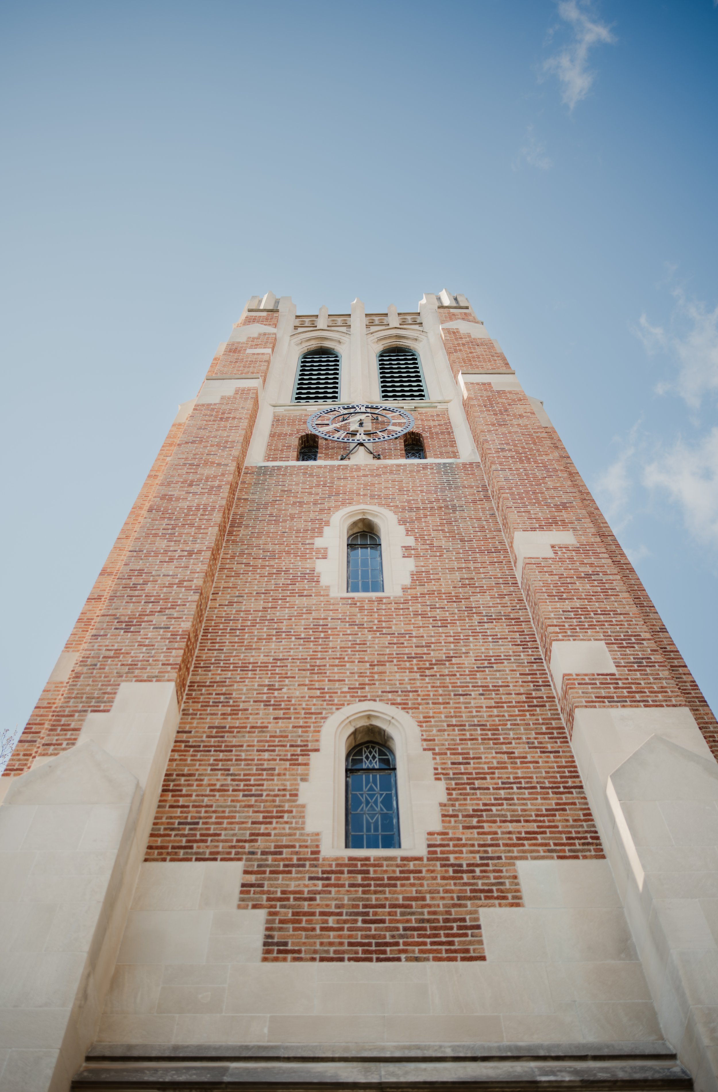 View of a tall brick church steeple with clock, set against a blue sky with light clouds.
