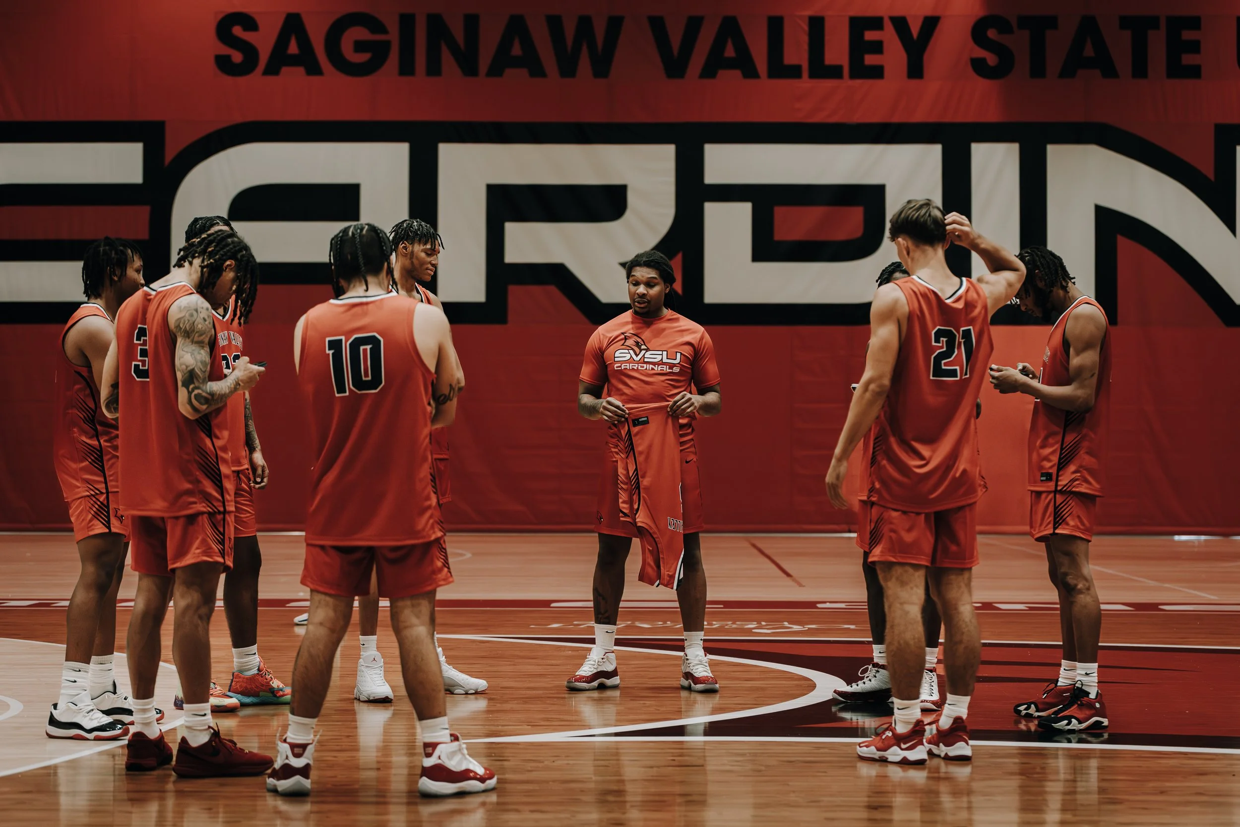 A women's basketball team in red uniforms gathered around a coach or team leader at Saginaw Valley State University in a gymnasium.