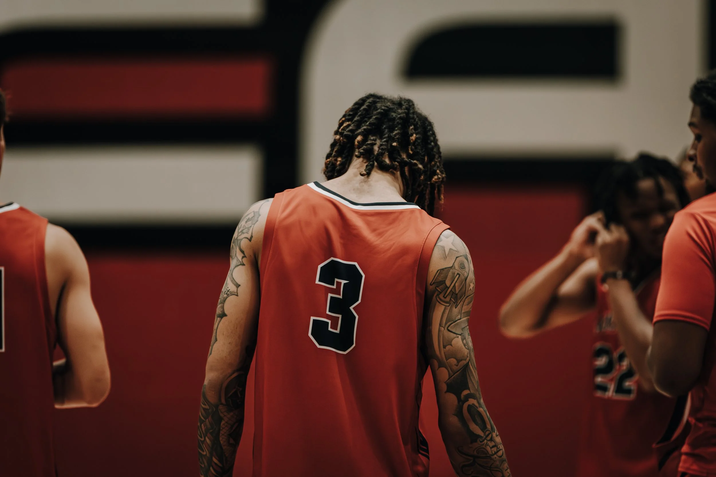 A group of female basketball players wearing red jerseys with black numbers, standing on the court during a game or practice, focusing on one player with tattoos on her arms and dreadlocks.