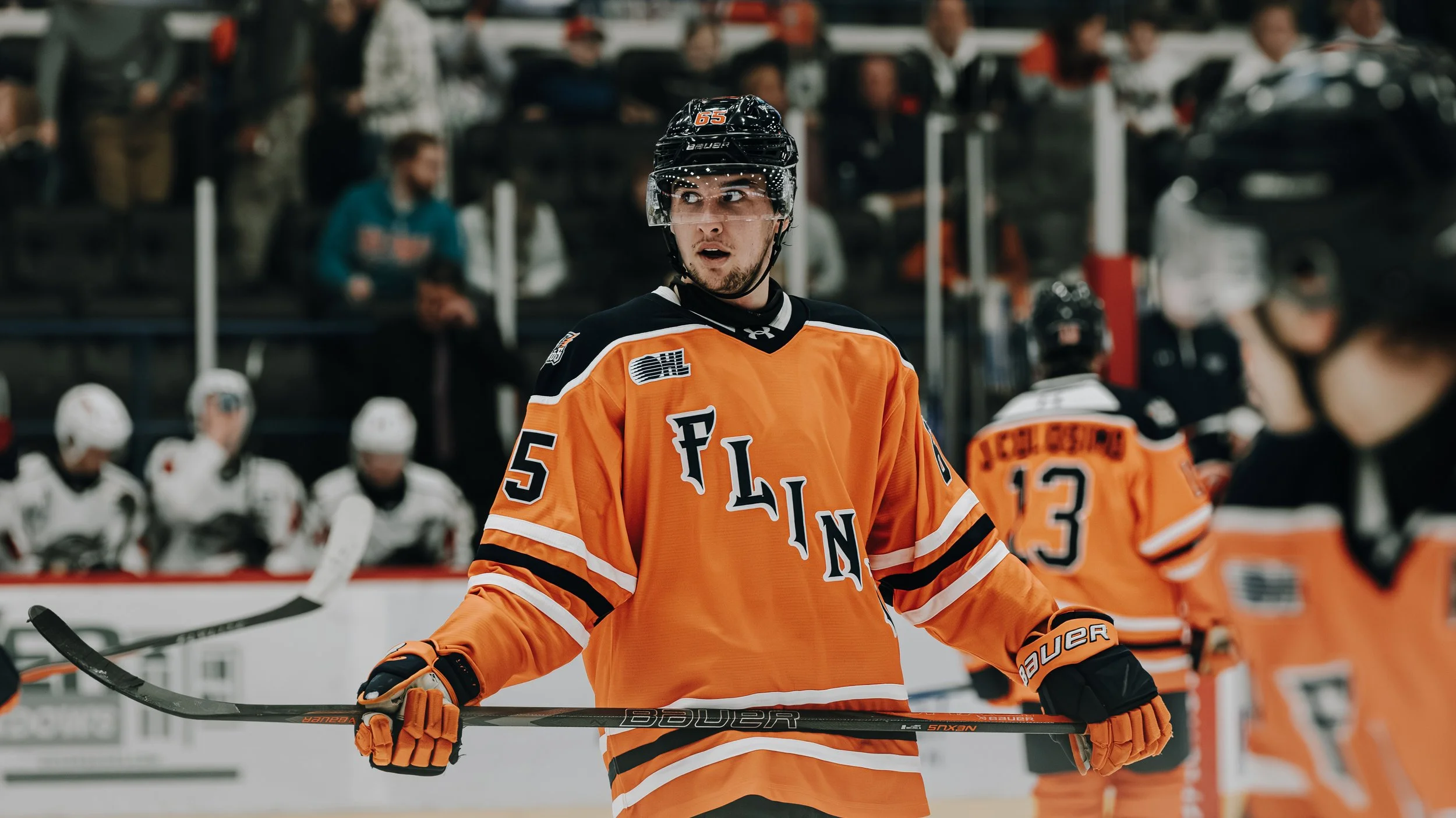 A hockey player wearing an orange New York Islanders jersey, #5, on the ice during a game, with other players and spectators in the background.