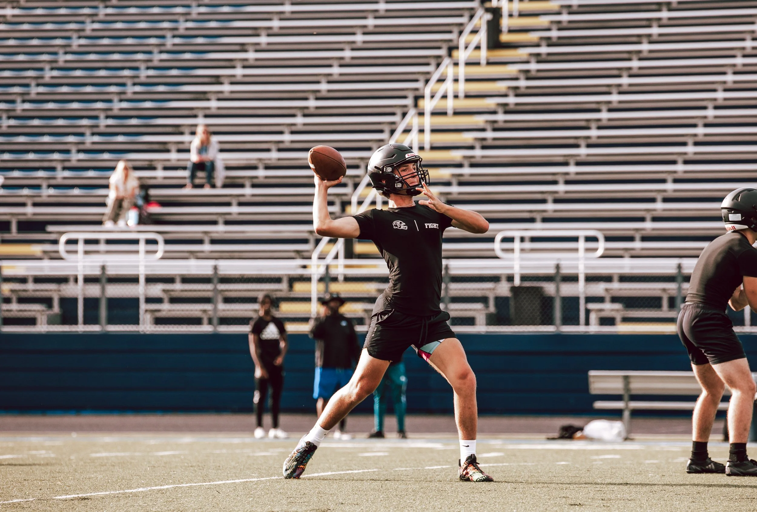 A person wearing a helmet is preparing to throw a football during a practice or game on a sports field, with empty bleachers in the background.