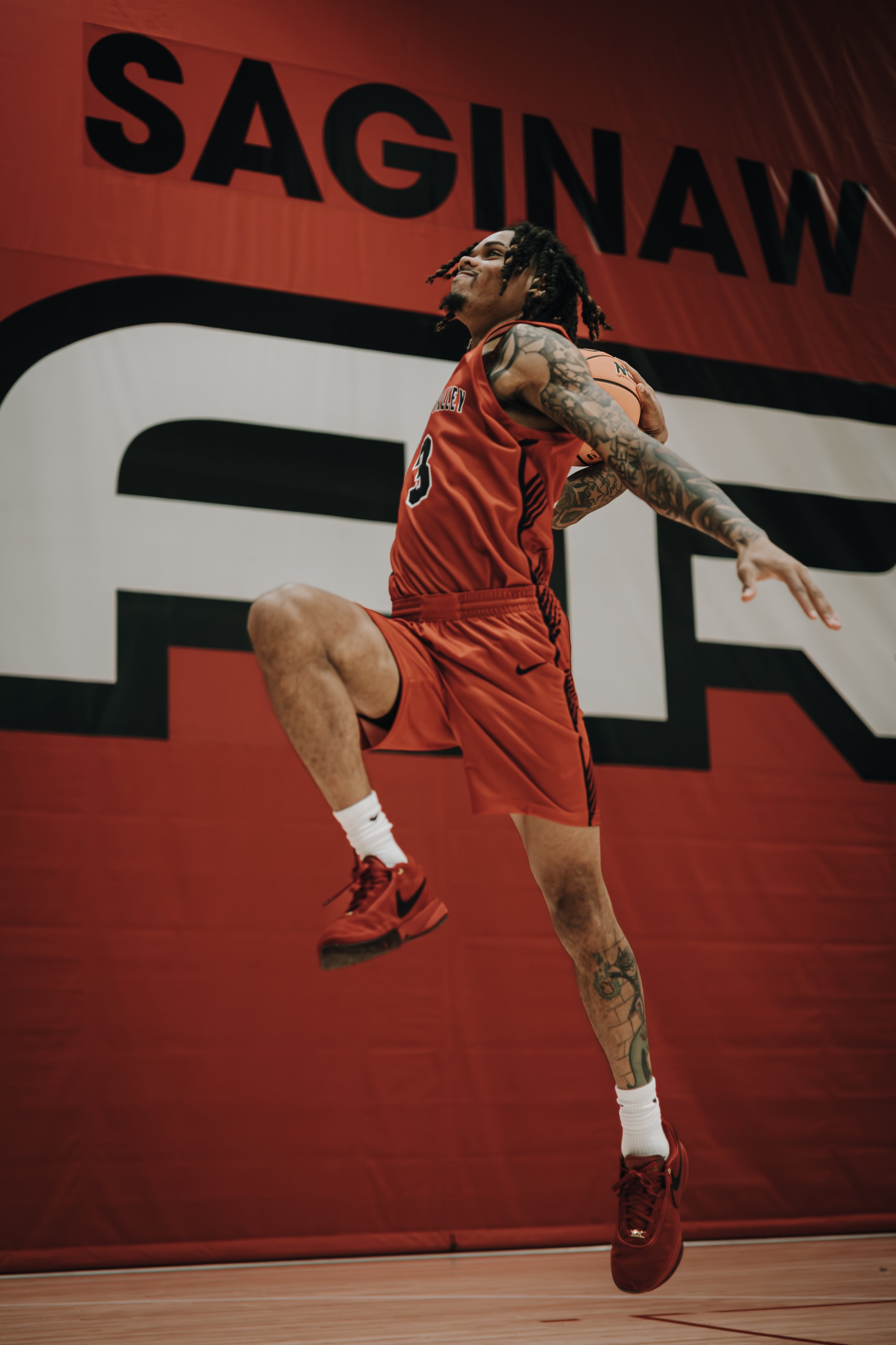 A basketball player in red uniform and sneakers jumping to shoot or dunk a basketball on an indoor court with a red wall and large black and white graphic in the background.