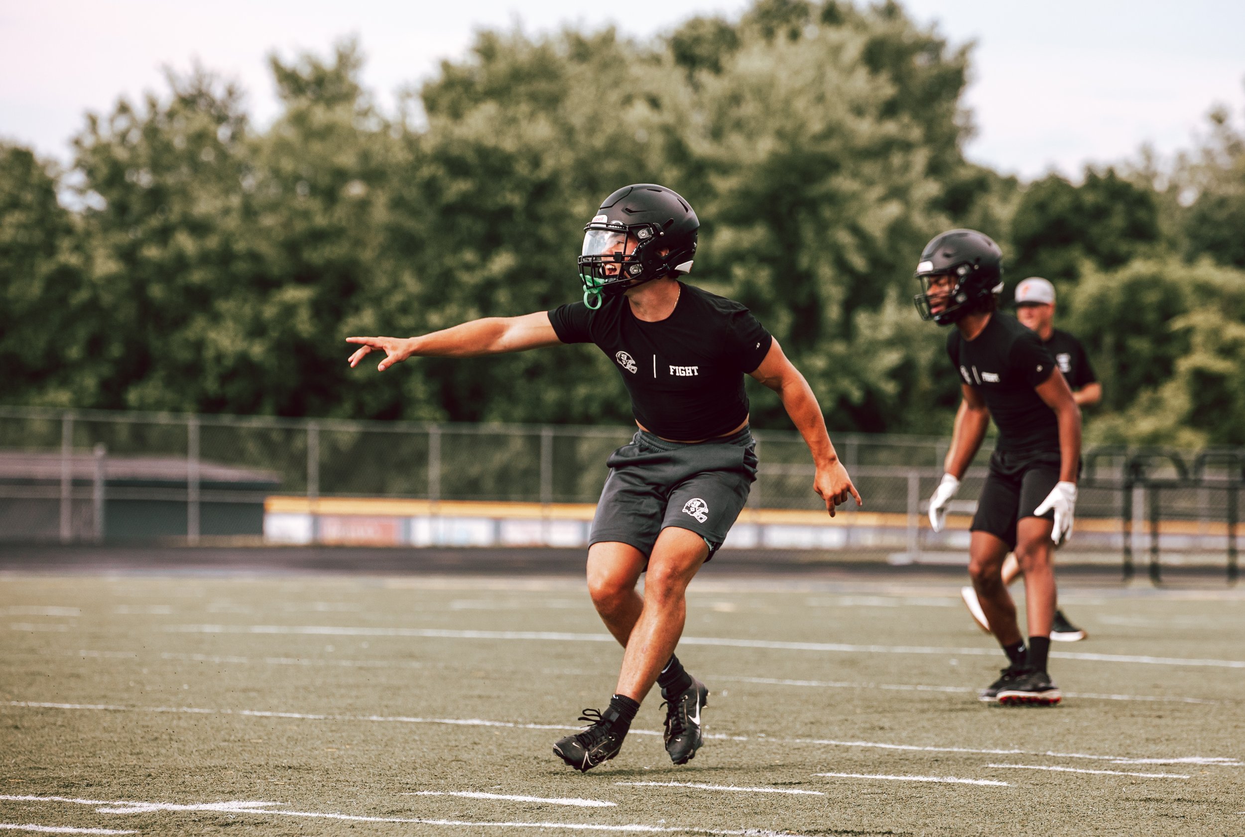 Two football players wearing helmets and black shirts practicing on an outdoor field, with one player pointing and the other looking on, during daylight.