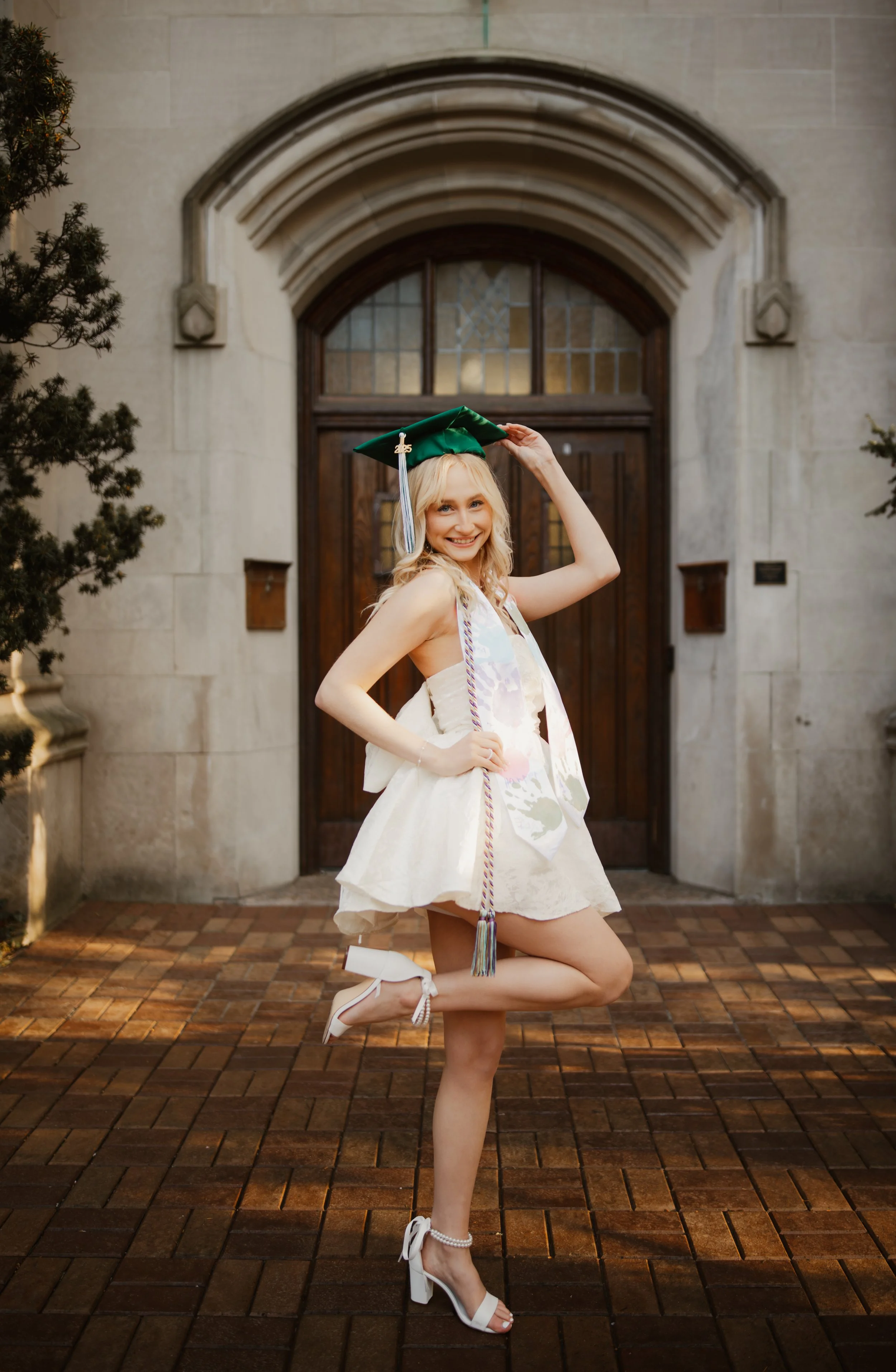 A young woman with blonde hair smiling, wearing a cream dress and white heels, poses with one knee on the ground while holding a green graduation cap in front of a stone building with an arched doorway, flanked by two trees.