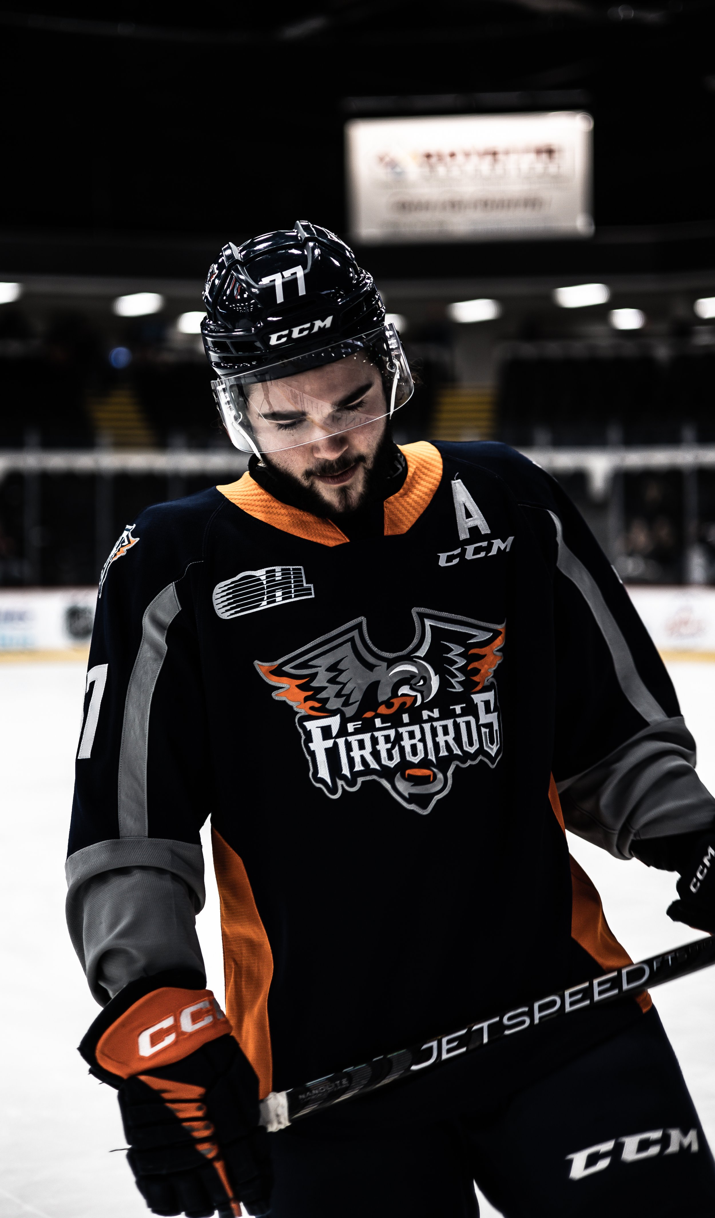 Hockey player in black and orange uniform with a logo that reads 'Ontario Firebirds,' wearing a helmet and holding a hockey stick on ice rink.