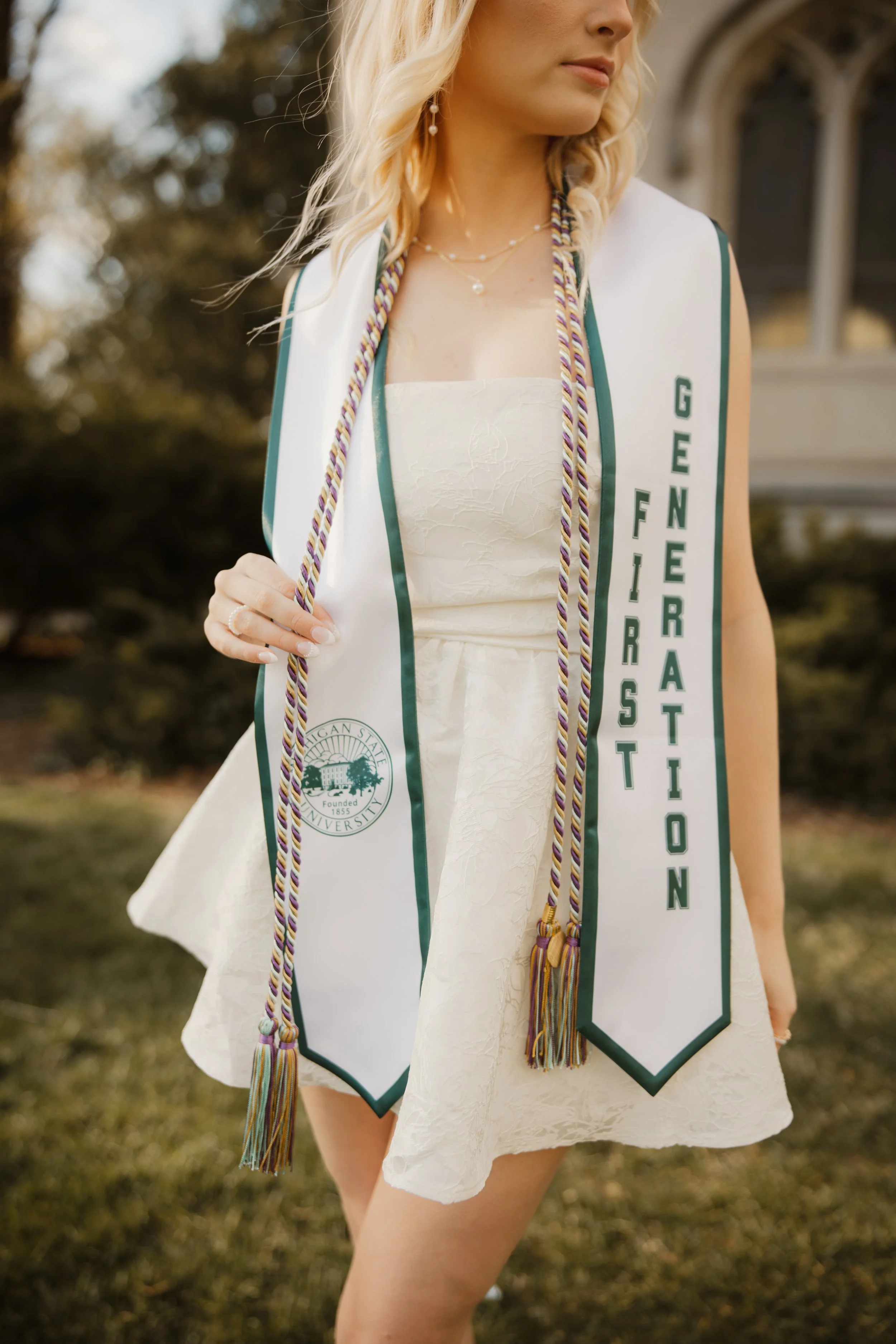 A young woman wearing a white dress and academic stoles with a graduation sash from Michigan State University, holding colorful cords, standing outdoors with trees and a building in the background.