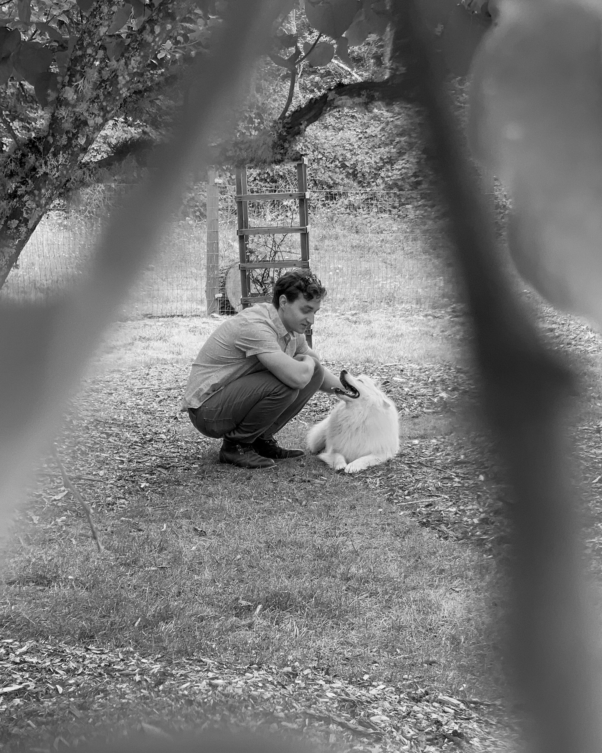 A young man crouching down and smiling at a fluffy, white dog on the grass in a park. The scene is viewed through a fence with trees and a wooden ladder in the background.