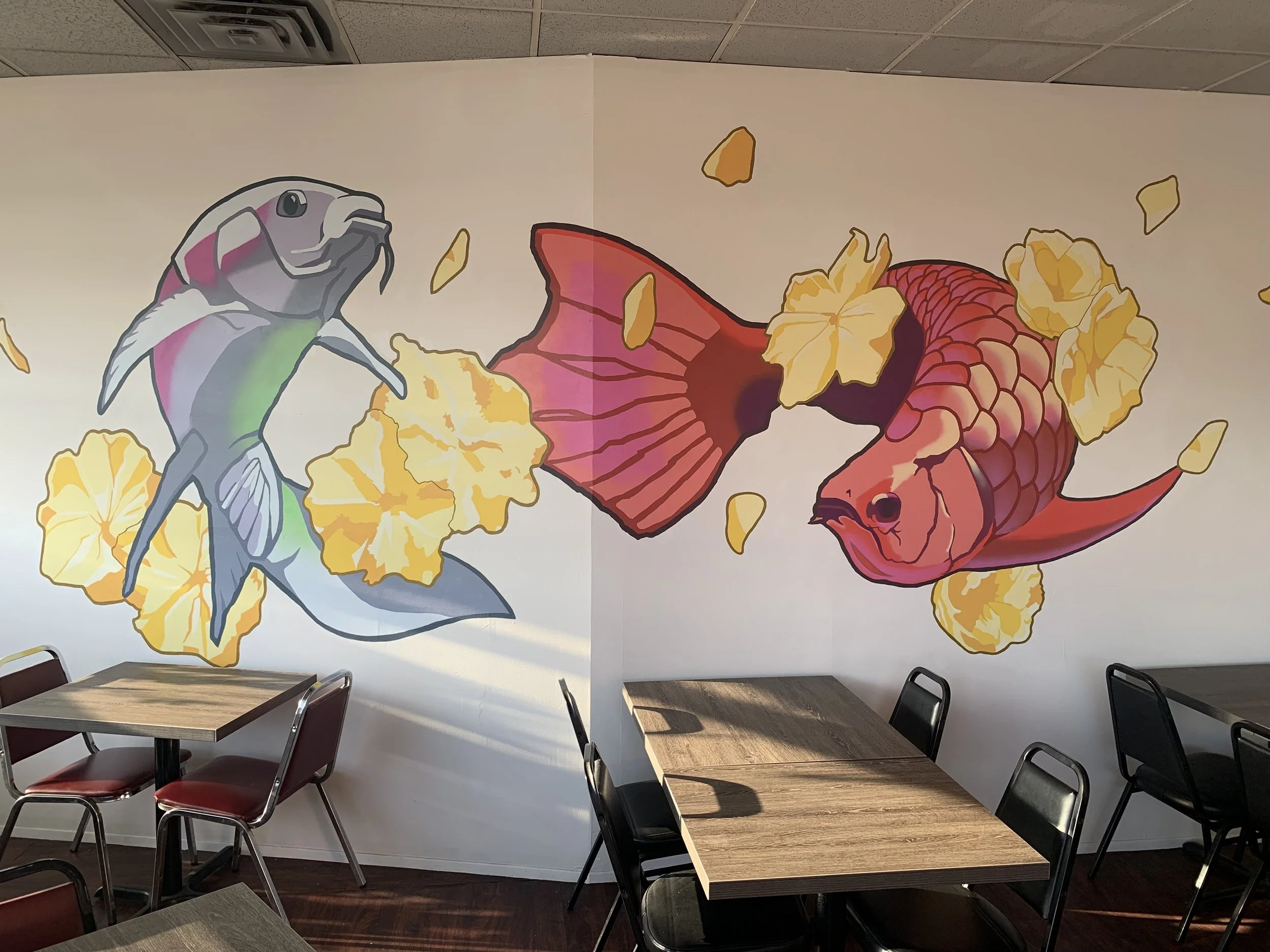 Indoor restaurant or cafeteria with a painted mural of a hummingbird and a fish on the wall. The mural features yellow flowers and pinkish-red leaves. Several tables and chairs are in the foreground.