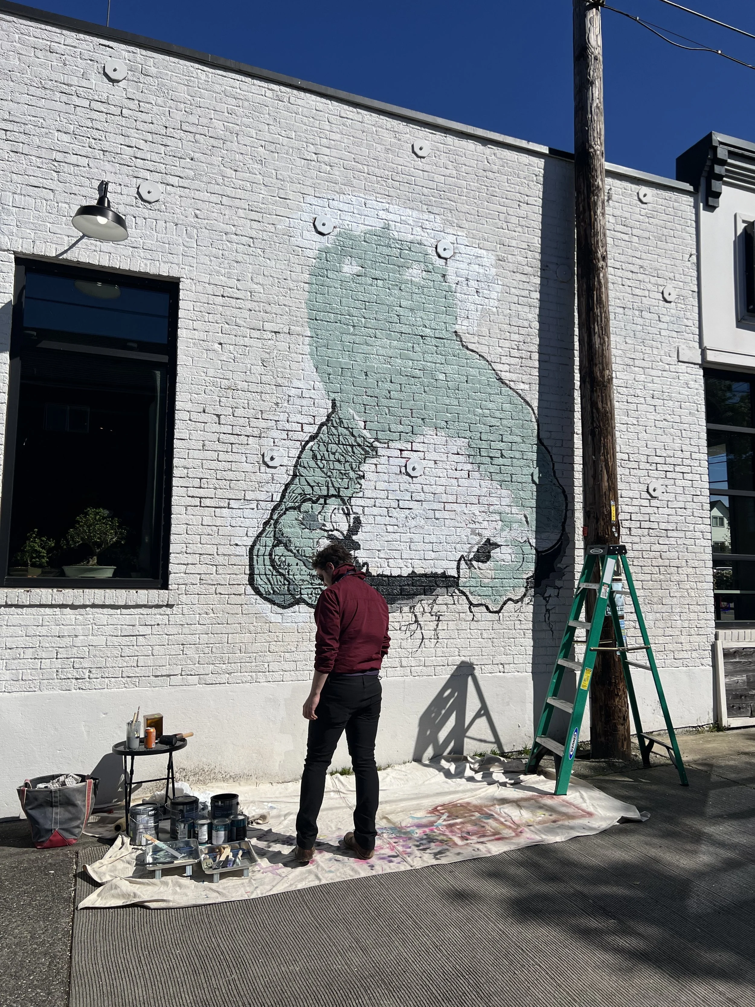 A person with dark hair and glasses wearing a maroon jacket painting a large mural of a silhouette of a female figure on a white brick wall. The mural shows the woman holding a paintbrush with a rainbow around her head. Art supplies, cans of paint, and brushes are on the ground covered with a white sheet. A ladder leans against a wooden utility pole nearby, and a small black table with painting tools is also present.