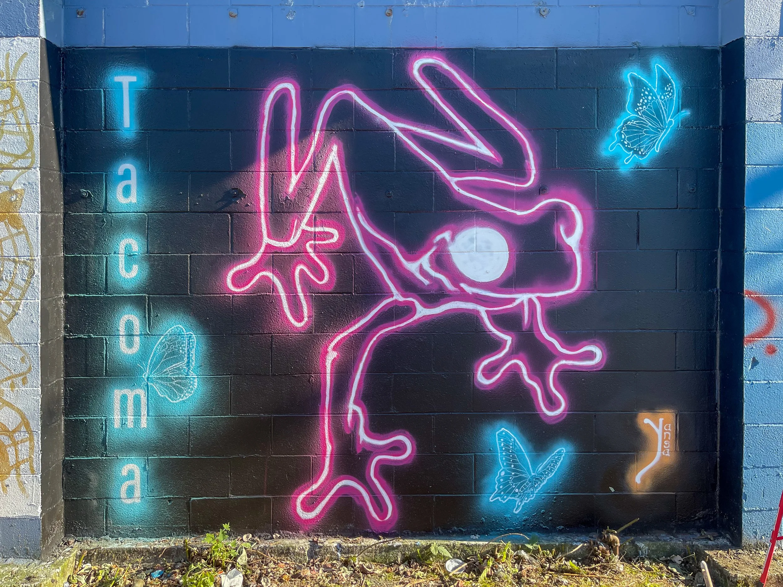 Neon graffiti art on a brick wall featuring a pink outline of a human figure in a yoga pose with one hand touching the ground and the other reaching upward, surrounded by blue neon butterflies and the word 'peace' spelled vertically in blue neon letters.