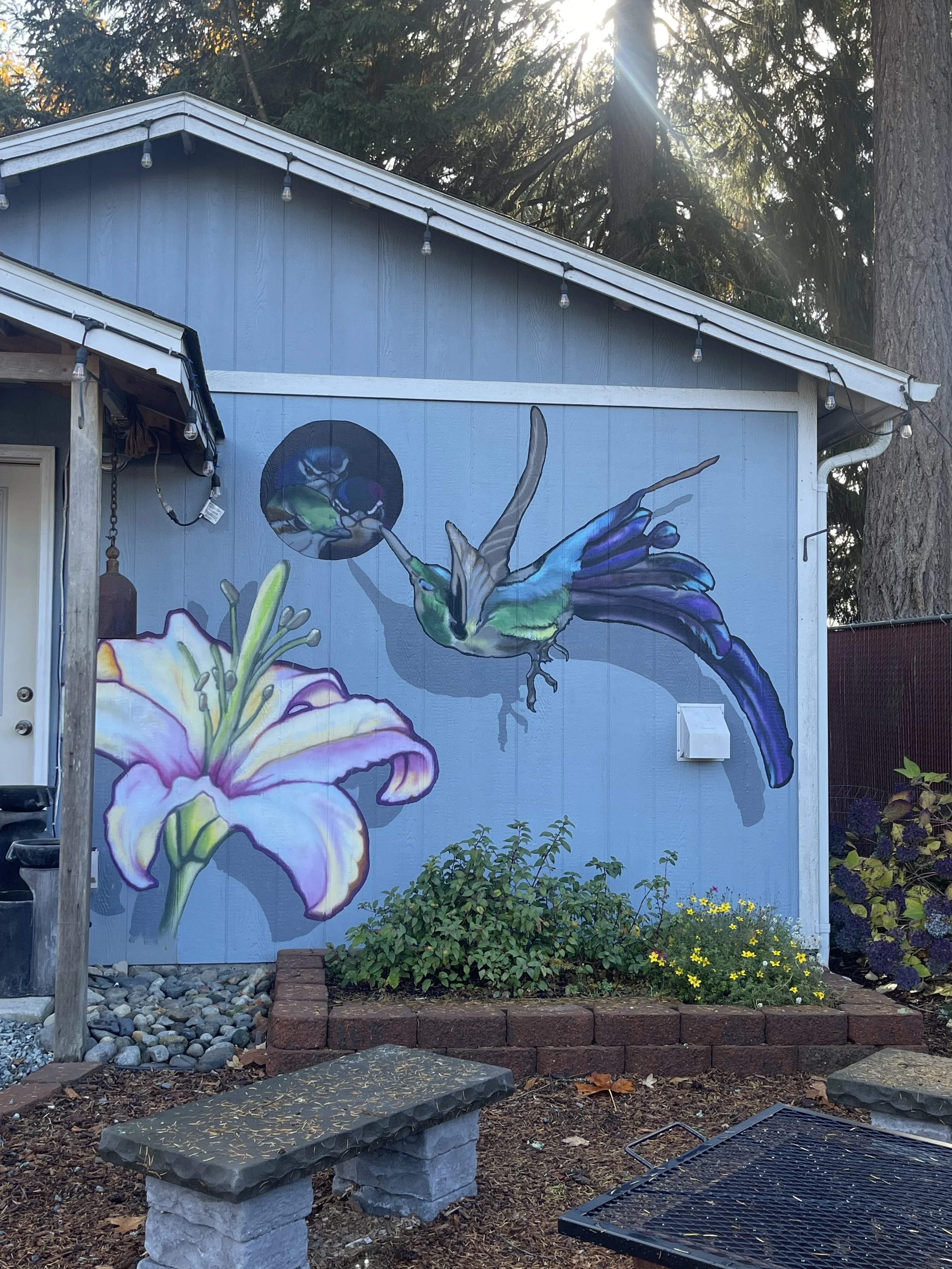A mural of a hummingbird hovering beside a large lily flower on the side of a house, with a garden and a stone bench in the foreground.
