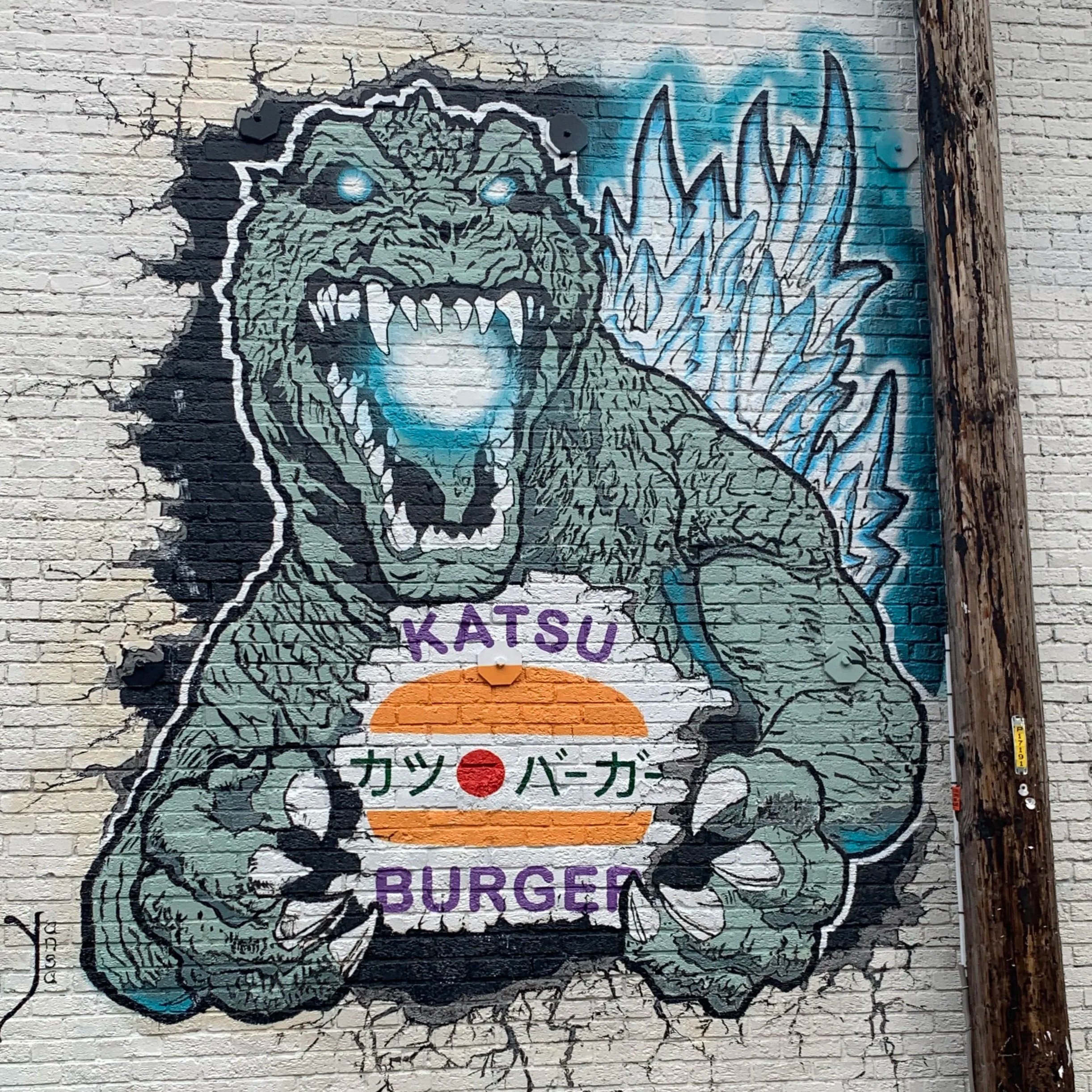 Street art graffiti of a dinosaur with blue eyes, sharp teeth, and spikes along its back, on a brick wall adjacent to a wooden utility pole. Text in Japanese and English is partially visible at the bottom.