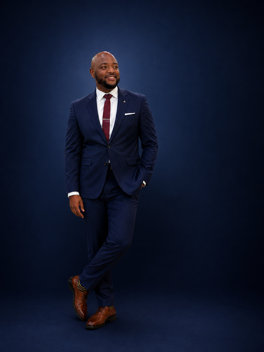 A man in a navy blue suit, white shirt, maroon tie, and brown dress shoes standing against a dark blue background. He has a bald head and beard, smiling with one hand in his pocket.