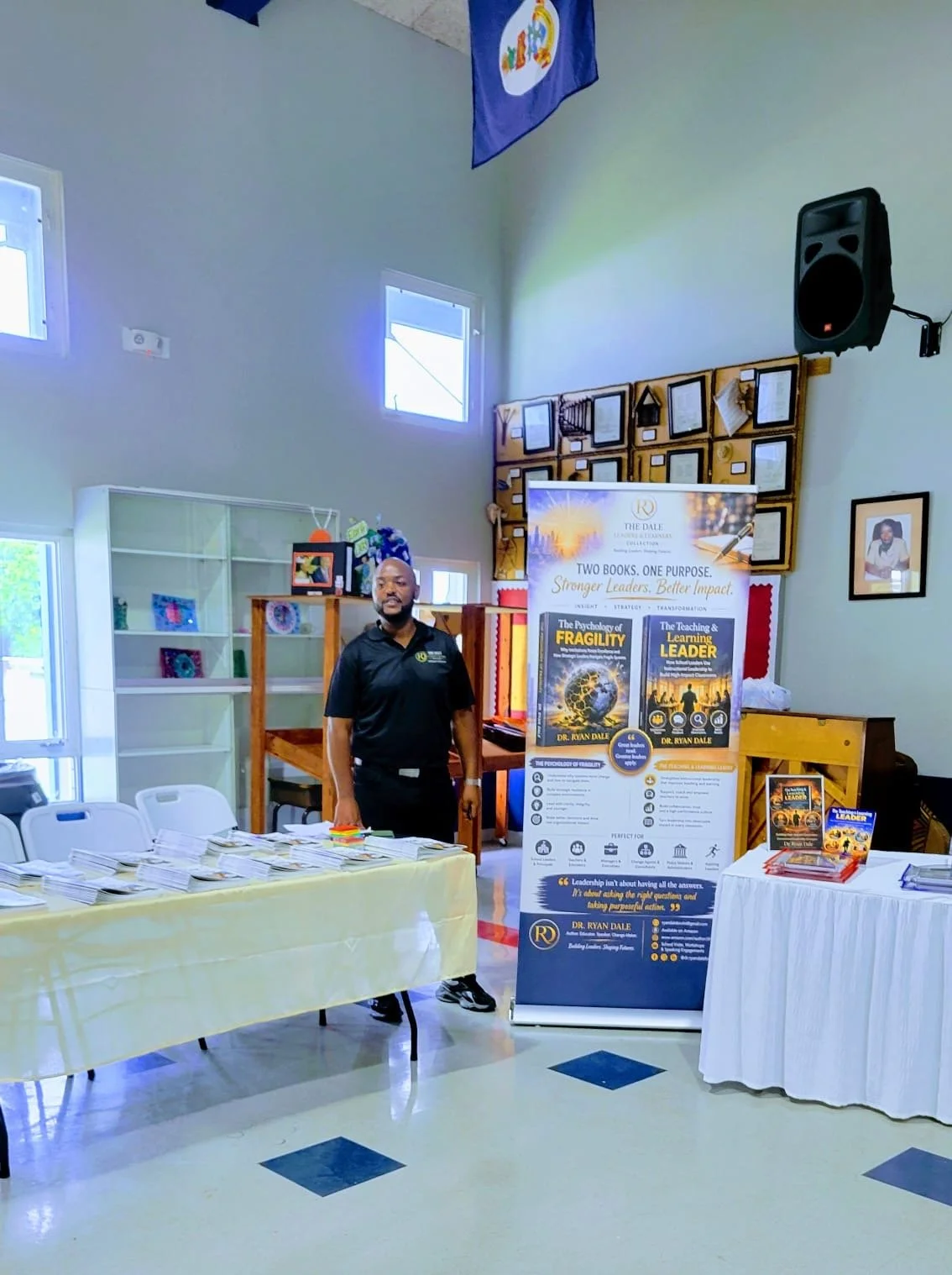A man stands next to a booth with a sign promoting two books about leadership and psychology. The booth has a table with papers and a display of books, with a framed photo on the wall and a speaker mounted on the wall in a brightly lit room.