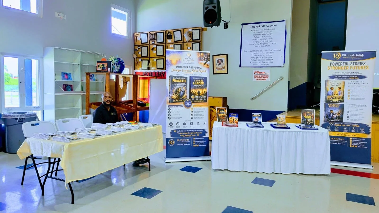 An indoor event space with tables displaying books and promotional materials for Dr. Ryan Dale. A man is sitting at a table on the left, and there are banners and posters promoting books and leadership topics in the center and right. The room is well-lit with natural light coming from windows, and there are framed pictures and a poem on the wall.