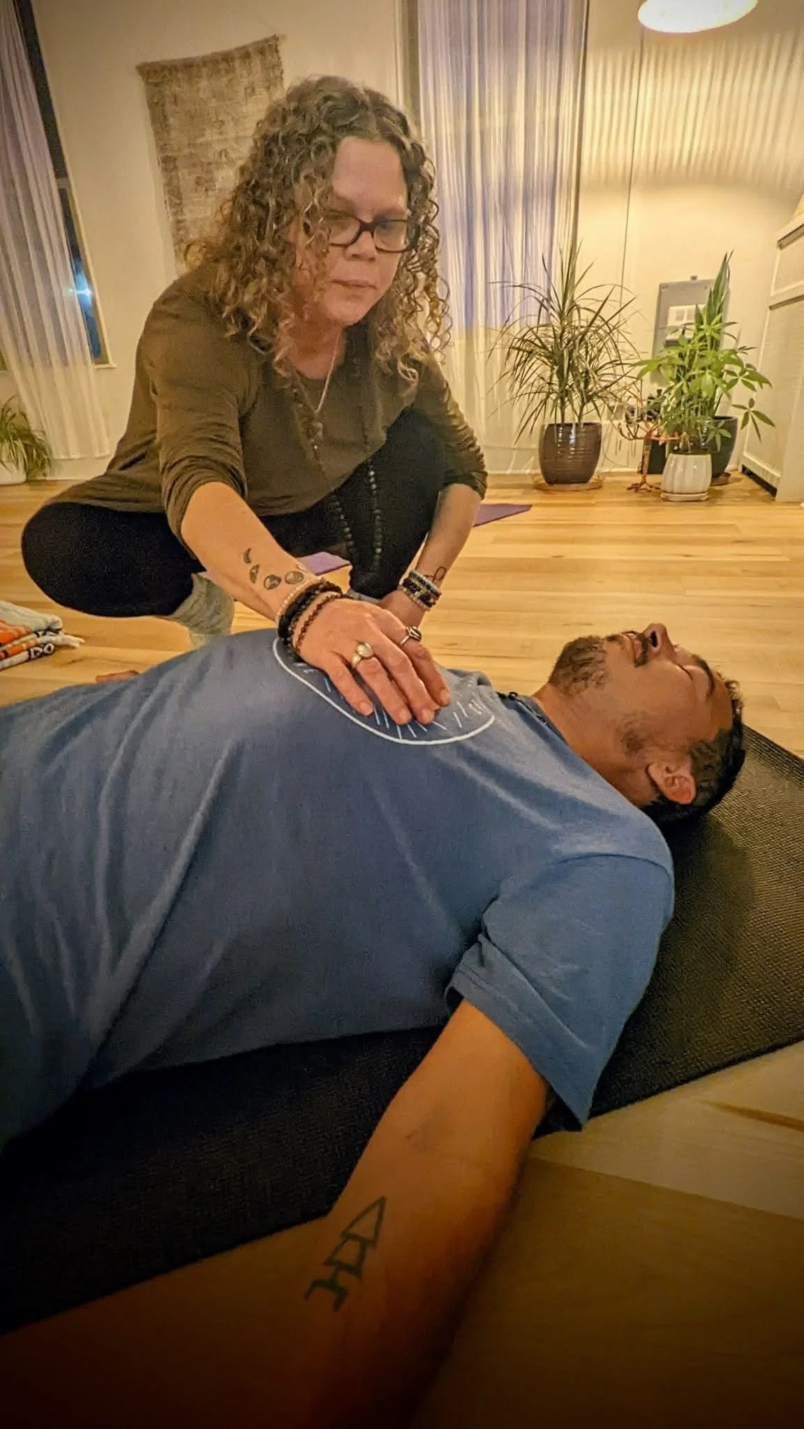 A woman with curly hair and glasses performing reiki on a man lying on the floor, with her hand over his chest in a living room with plants and curtains.