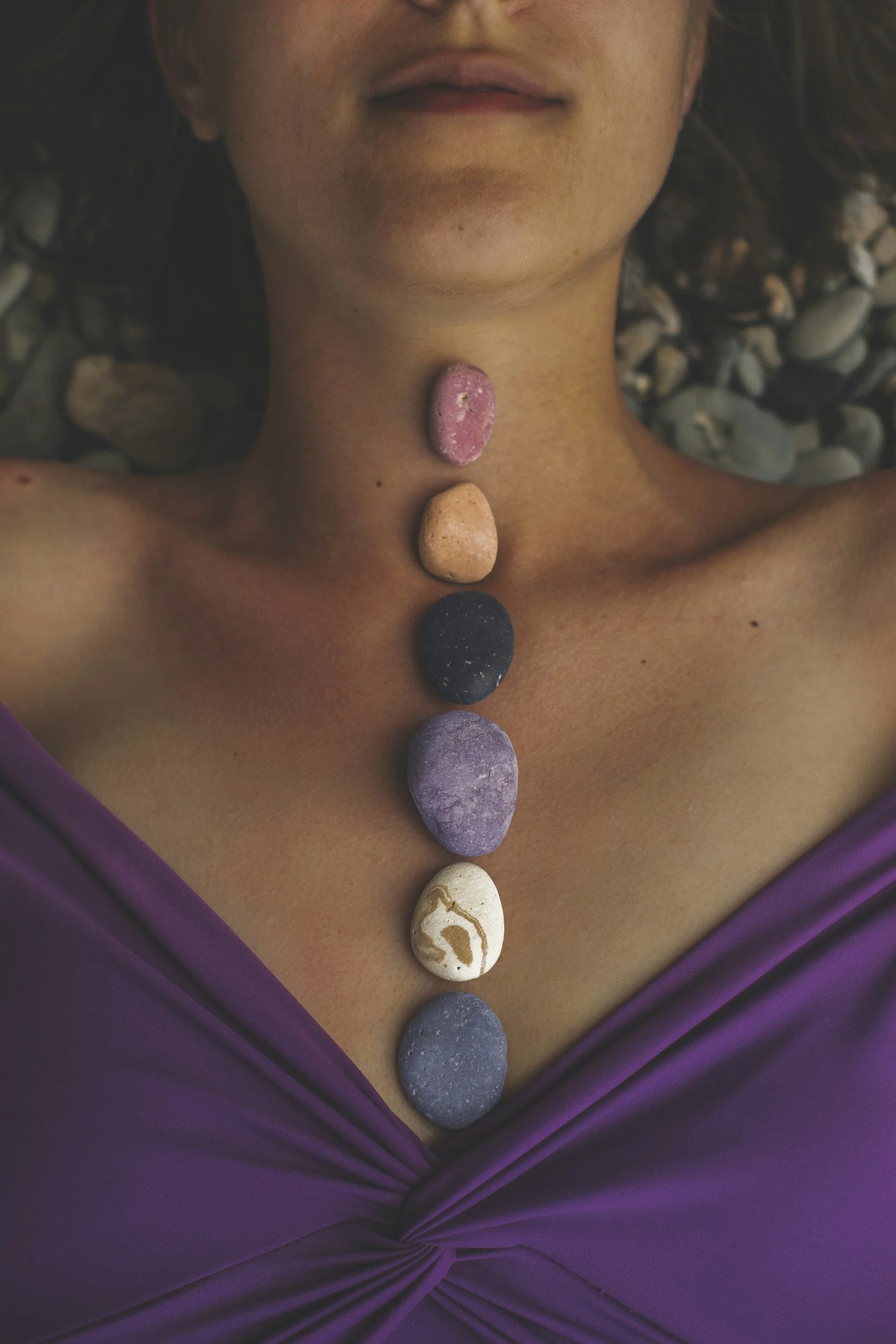 A person with a purple dress lying down on rocks, with seven colorful stones placed along their neck and chest, resembling a necklace.