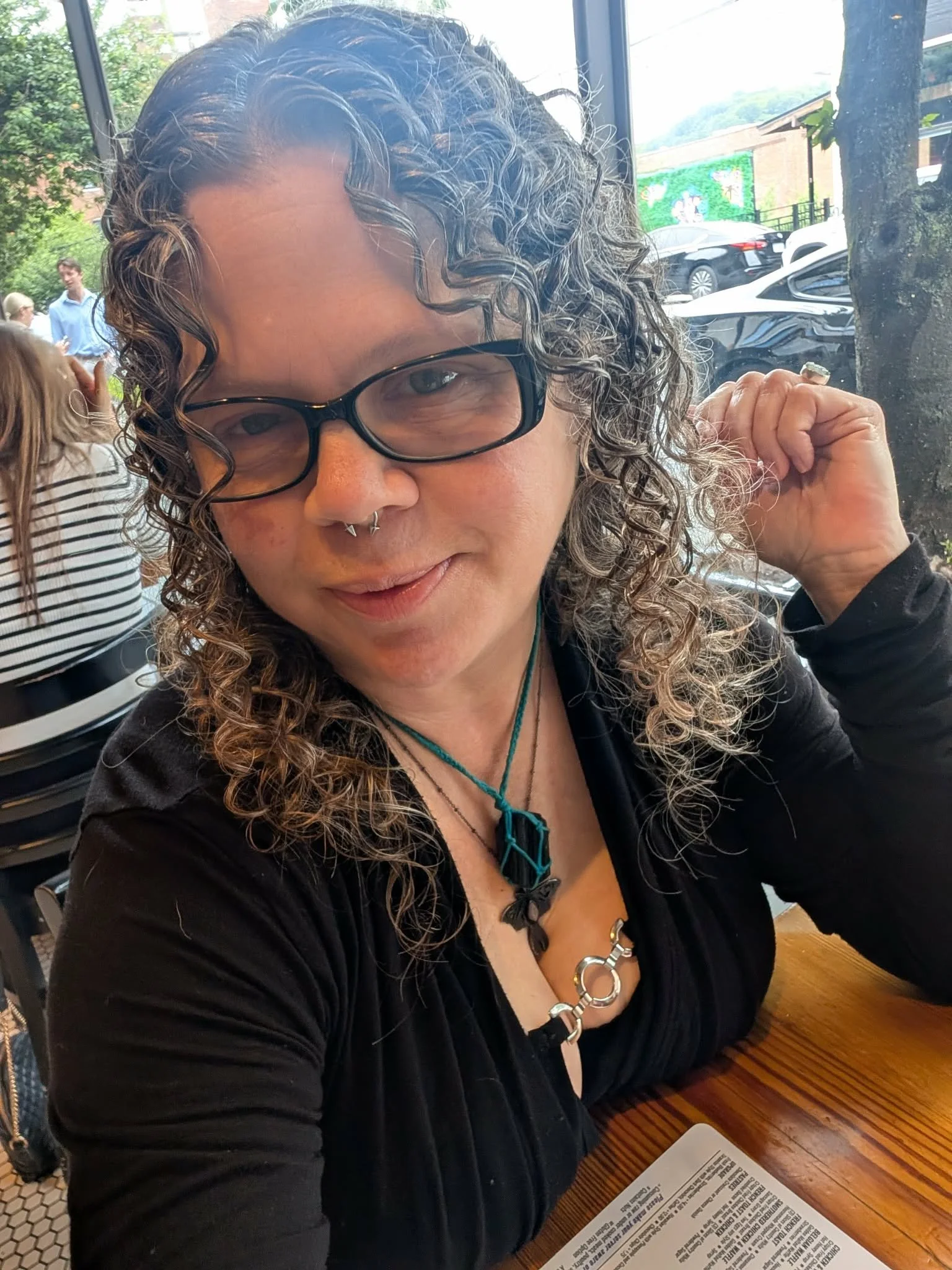 A woman with curly hair, glasses, and a septum piercing, sitting at a wooden table in a restaurant or cafe, with a menu in front of her. She is wearing a black top and a turquoise necklace with a geometric pendant.