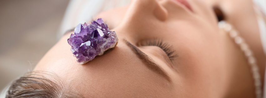Close-up of a woman lying down with a purple amethyst crystal on her forehead during a wellness or healing session.