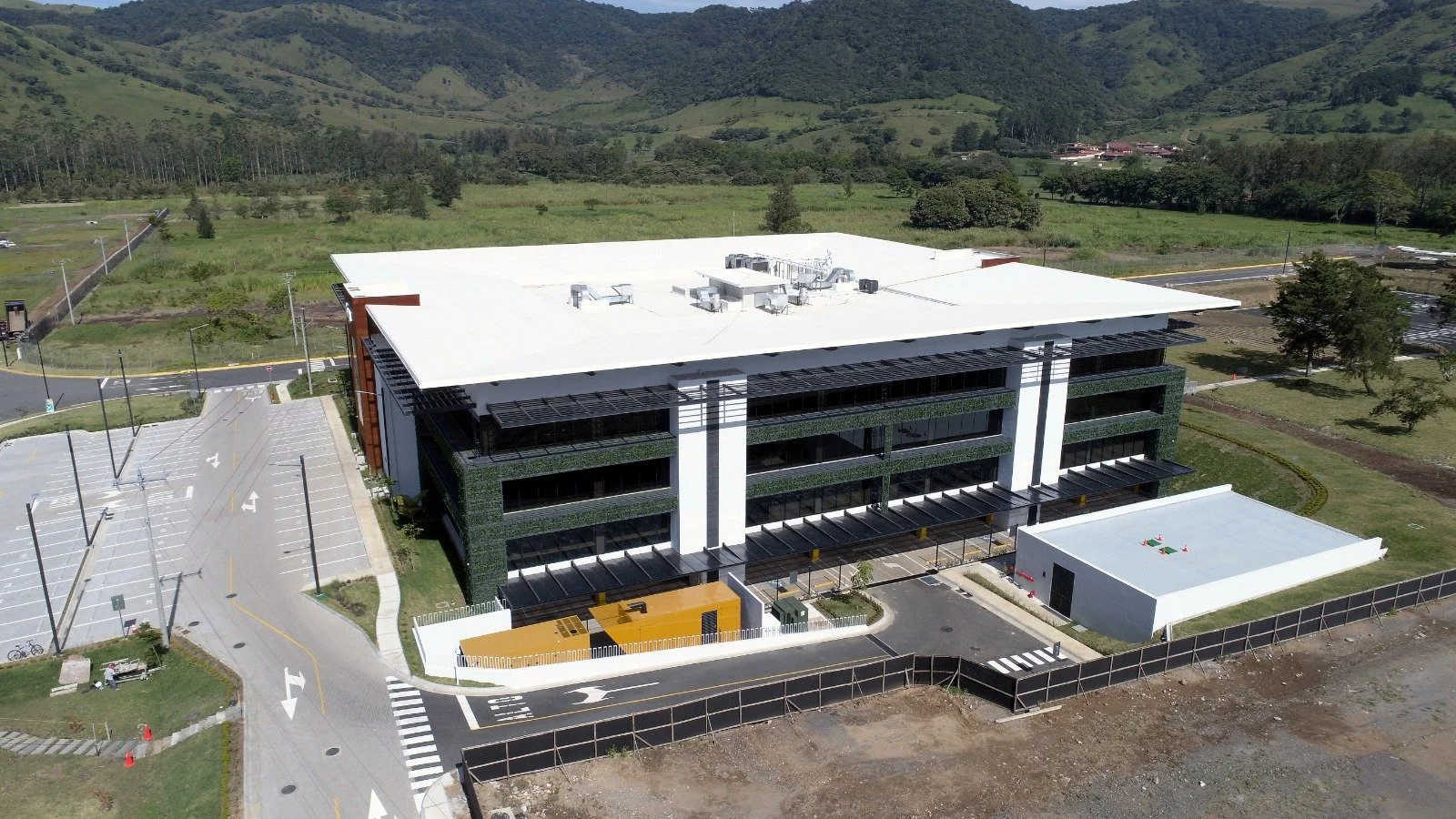 Edificio industrial con techo TPO de varios pisos con estacionamiento vacío, rodeado de áreas verdes y caminos, en un entorno natural con colinas y árboles.