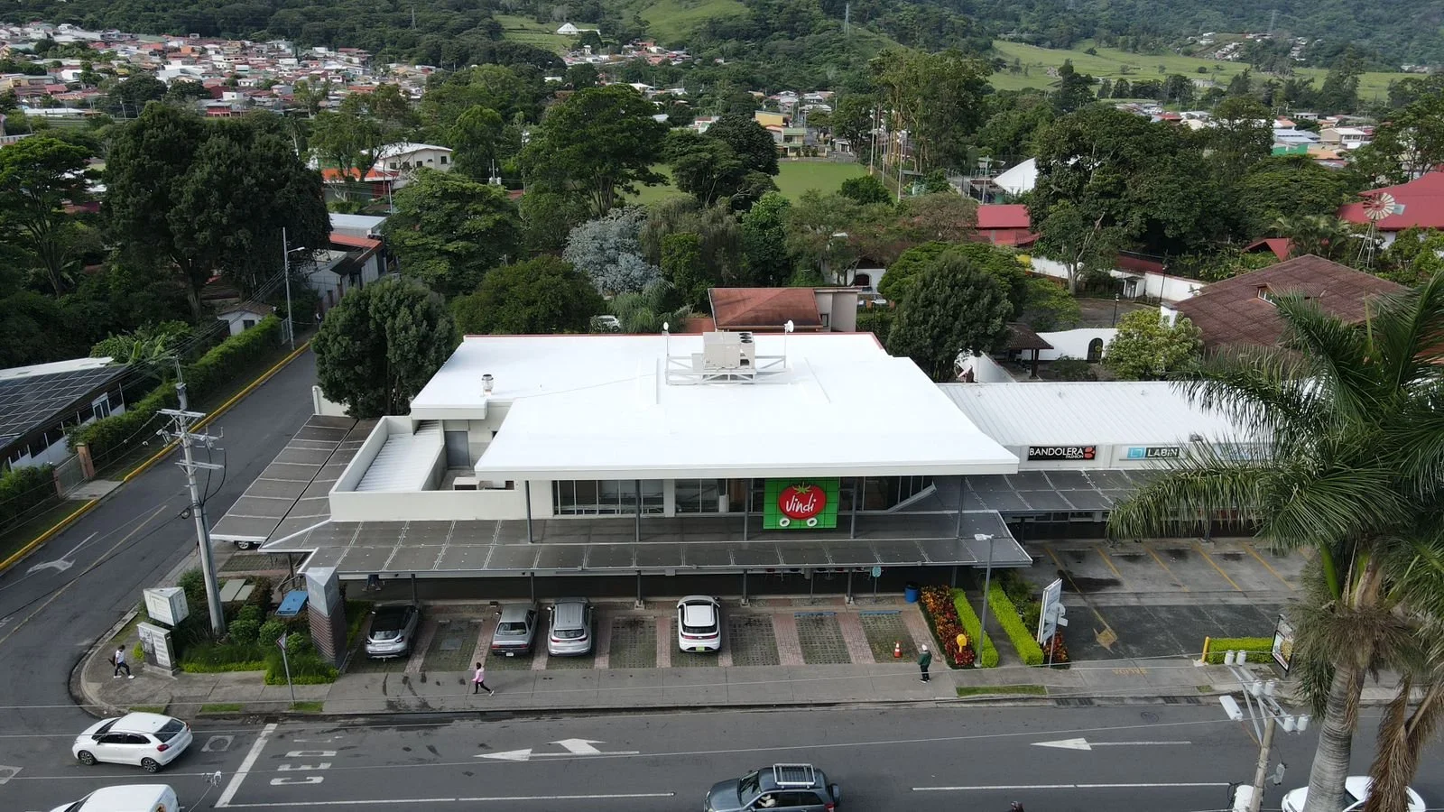 Vista aérea de un supermercado con retechado en membrana TPO. El letrero de 'Vindi' en la entrada, rodeado de árboles y una calle con vehículos y peatones.