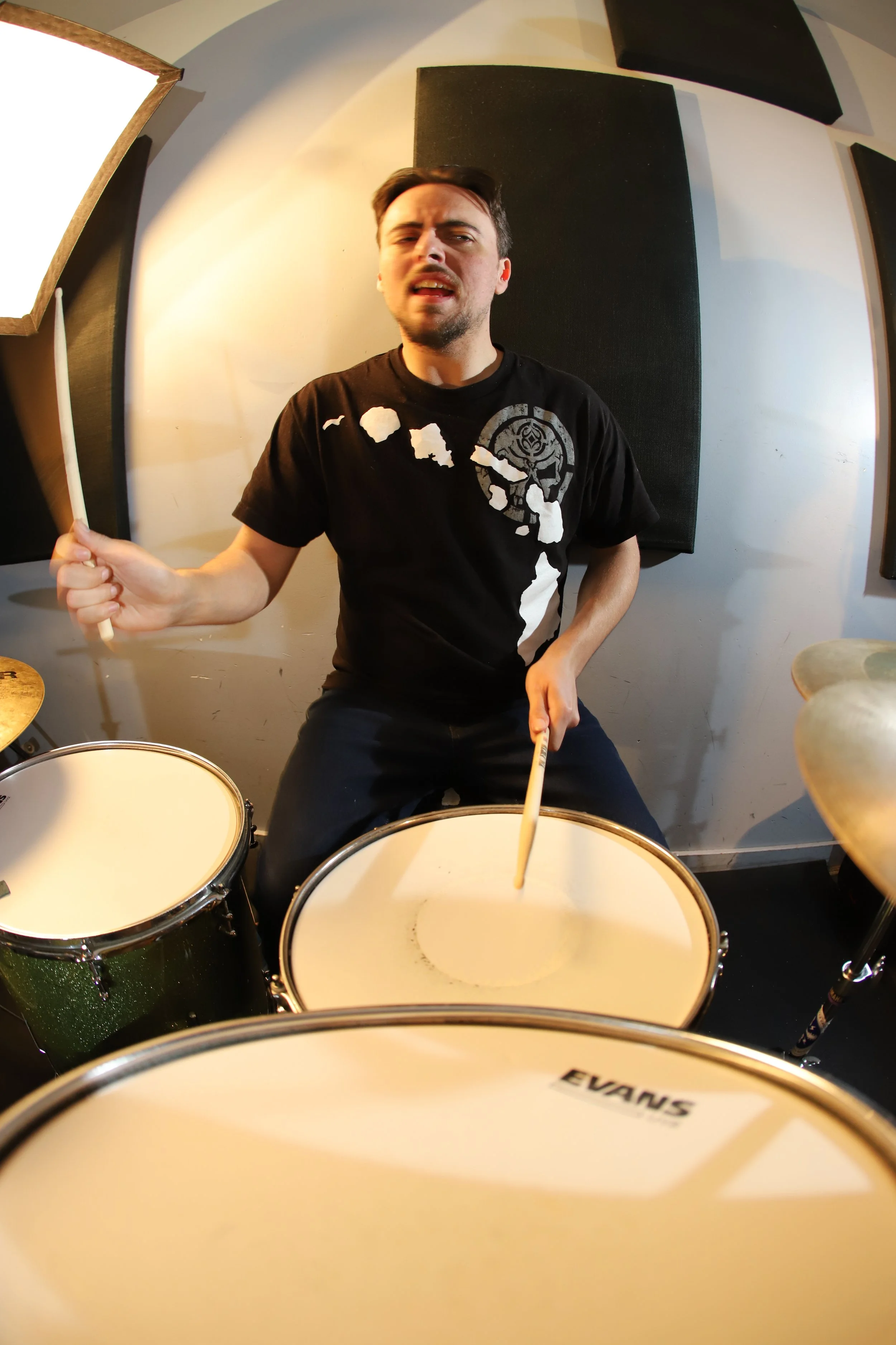 Jonny2Stix playing drums in a music studio, holding drumsticks, with black acoustic panels on the wall behind him.