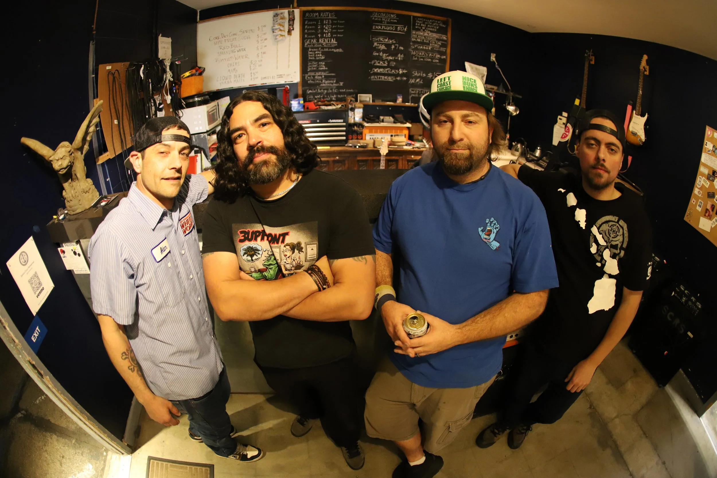Four men standing together in a room with dark walls, musical instruments on the wall, and a chalkboard menu in the background.