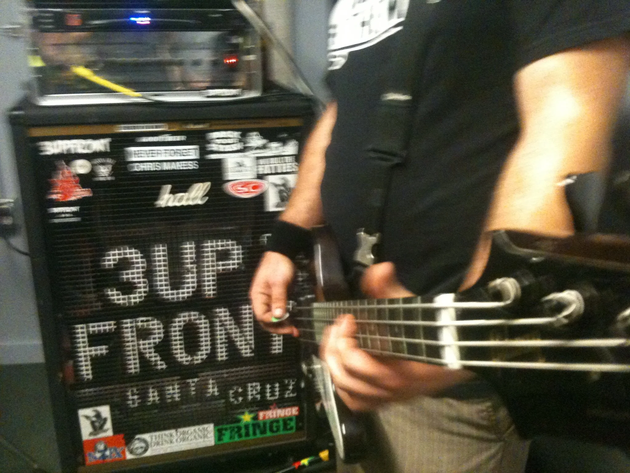 A person playing an electric bass guitar in front of a sign that reads 'SUP FRONT' with various stickers and artwork.