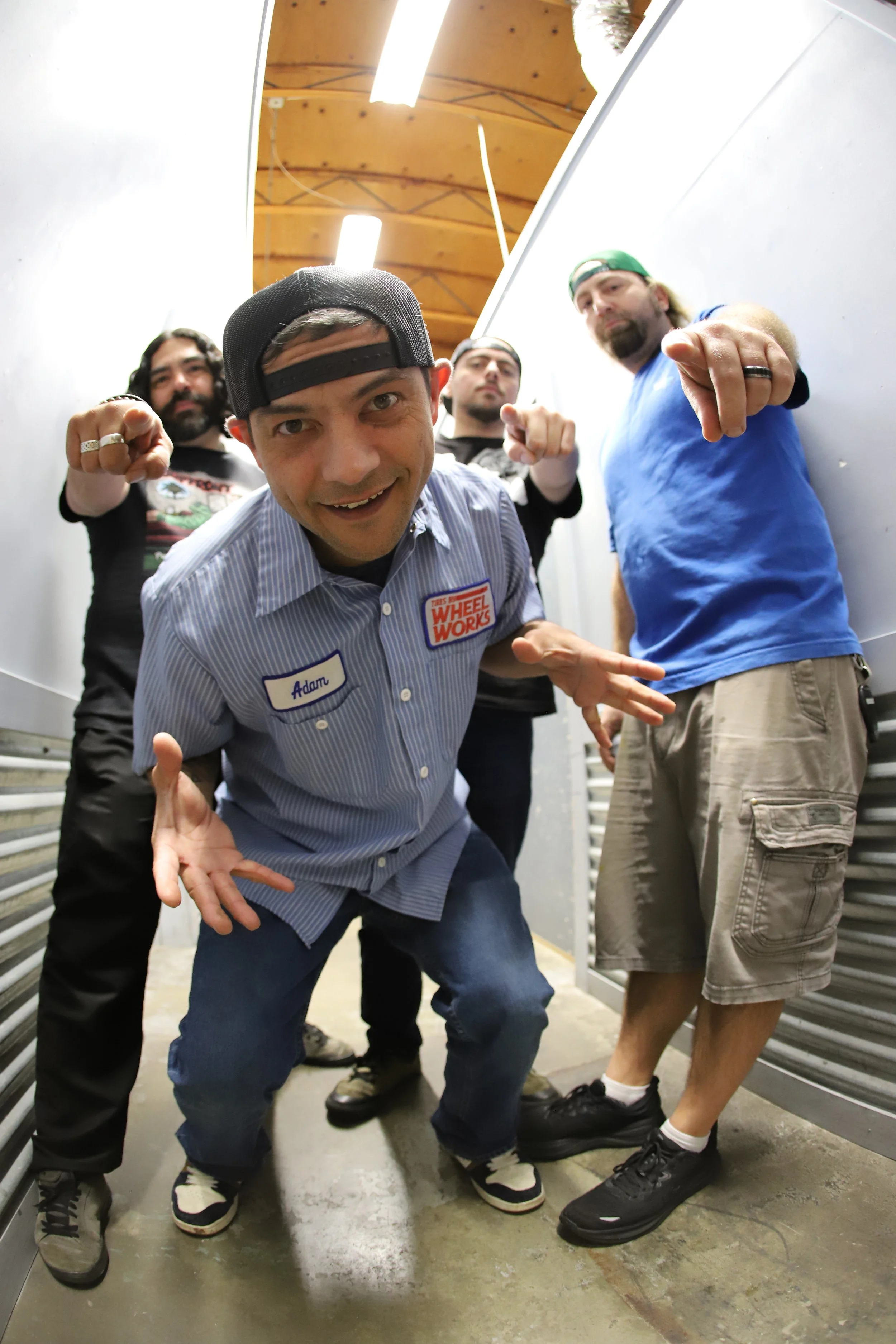 Four men standing in a narrow hallway, posing for the camera. The man in front is wearing a striped mechanic's shirt with a name tag that says 'Adam' and a patch that reads 'Wheel Works.' He is crouching slightly and smiling. The other three men are 